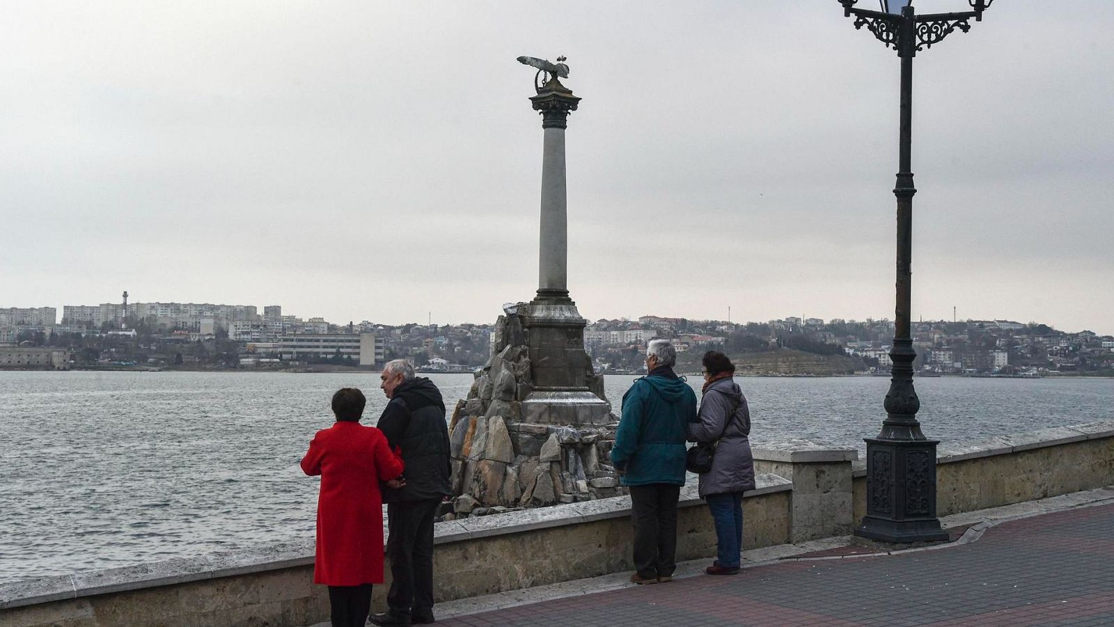 Monumento a los Barcos Hundidos en un día nublado, con cuatro personas paseando por un malecón y un paisaje urbano al fondo.