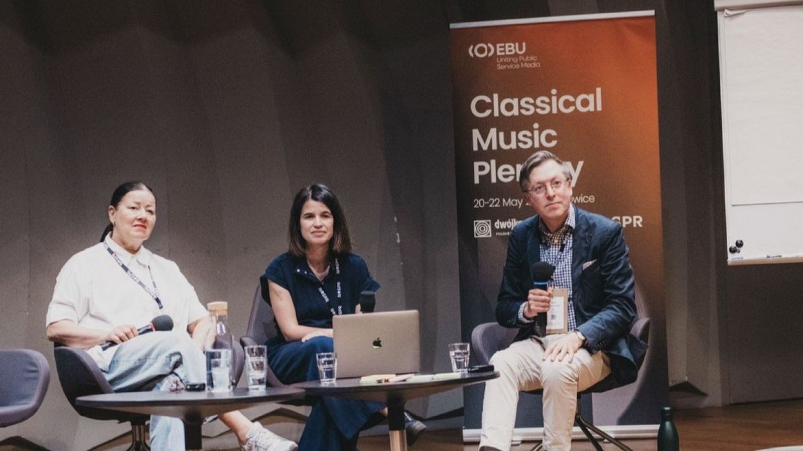 Panel de discusión "Classical Music Plenary" con tres oradores, un banner de EBU y logos de medios de comunicación. Se aprecian micrófonos, botellas de agua y un portátil sobre la mesa.