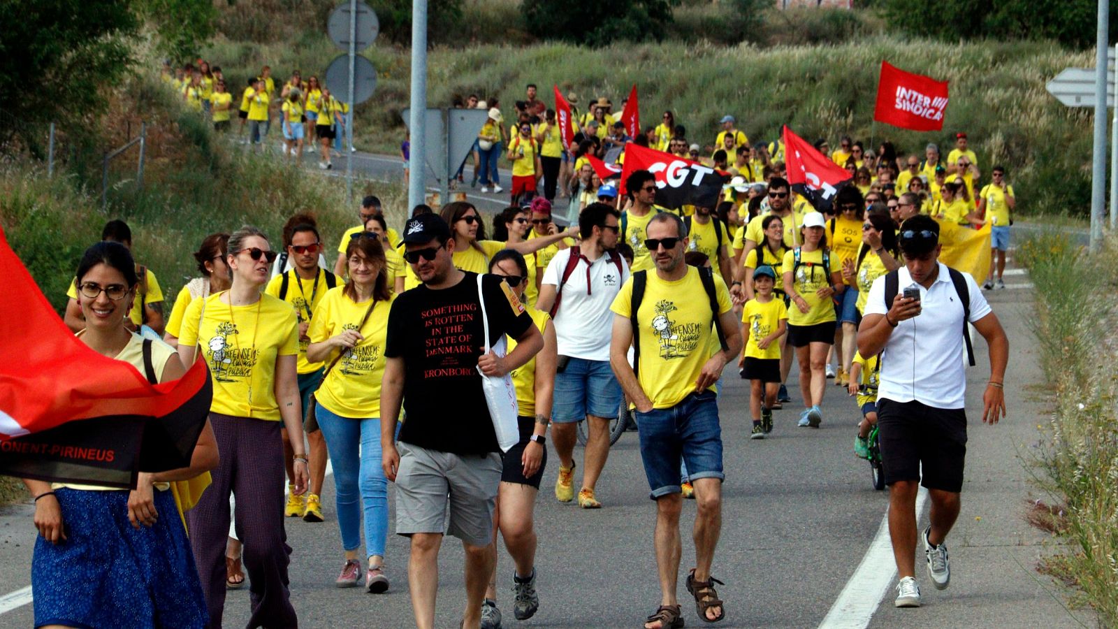 Manifestación con camisetas amarillas, banderas de "INTERSINDICAL" y "CGT", y mensajes de protesta en una carretera soleada.