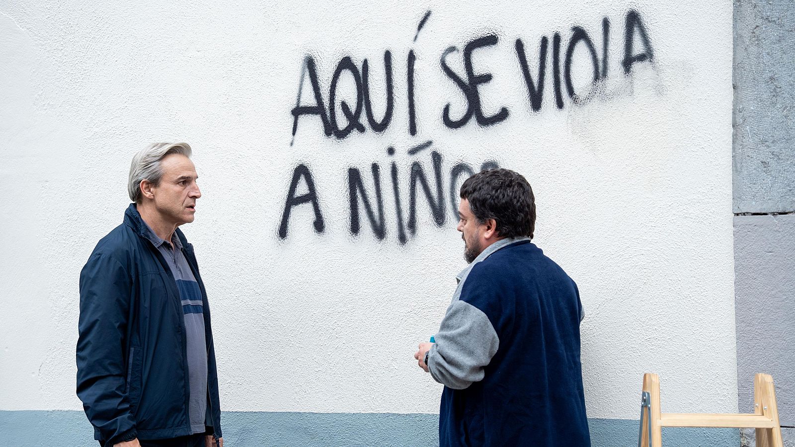 Javier Cámara y otro hombre observan un grafiti con un mensaje perturbador en una pared exterior. Se aprecia una escalera de madera en la parte inferior derecha.