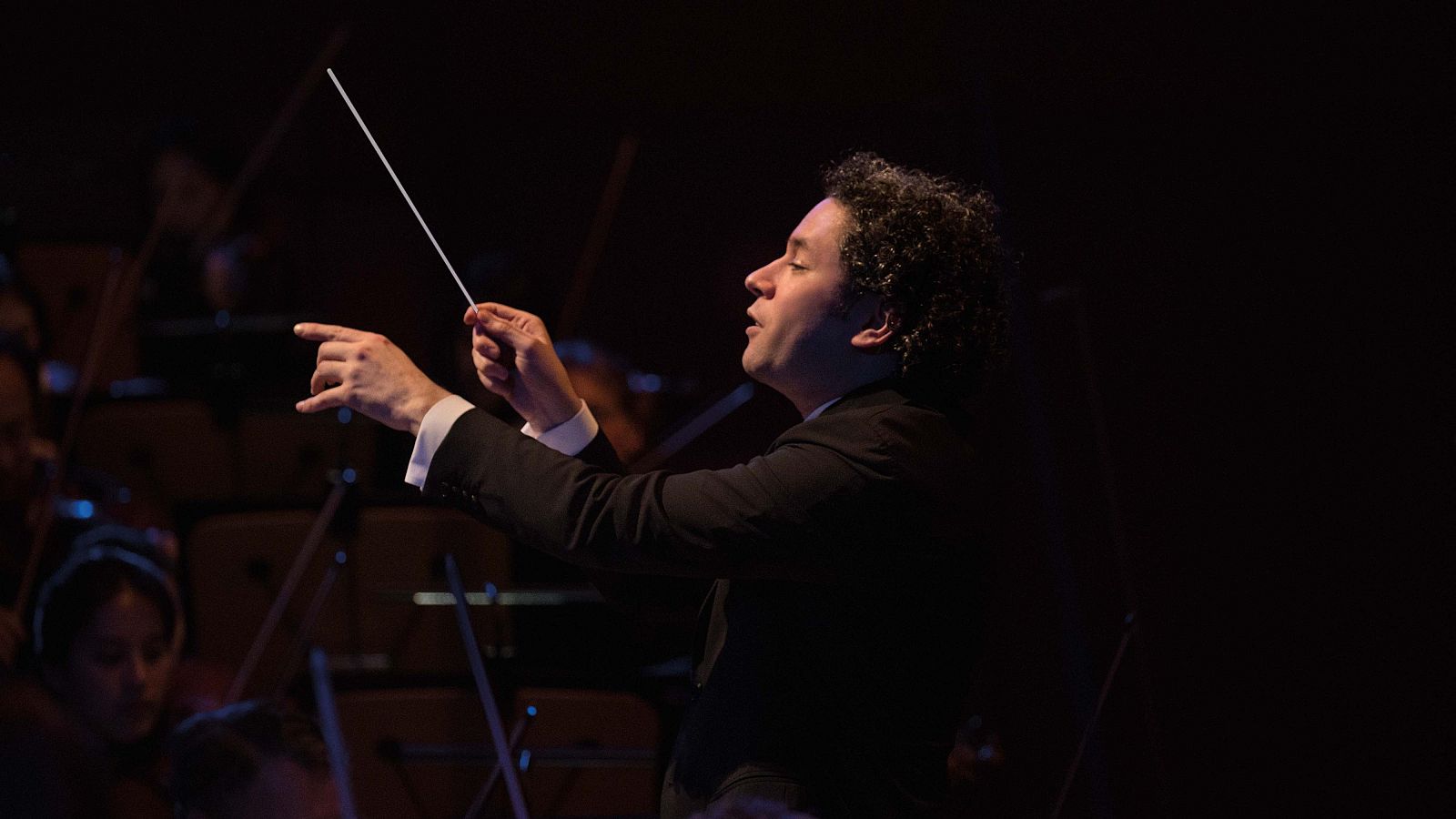 Gustavo Dudamel dirige con pasión, batuta en mano y dedo índice apuntando, en un concierto con orquesta. Su rostro muestra concentración y la chaqueta oscura contrasta con la camisa blanca.