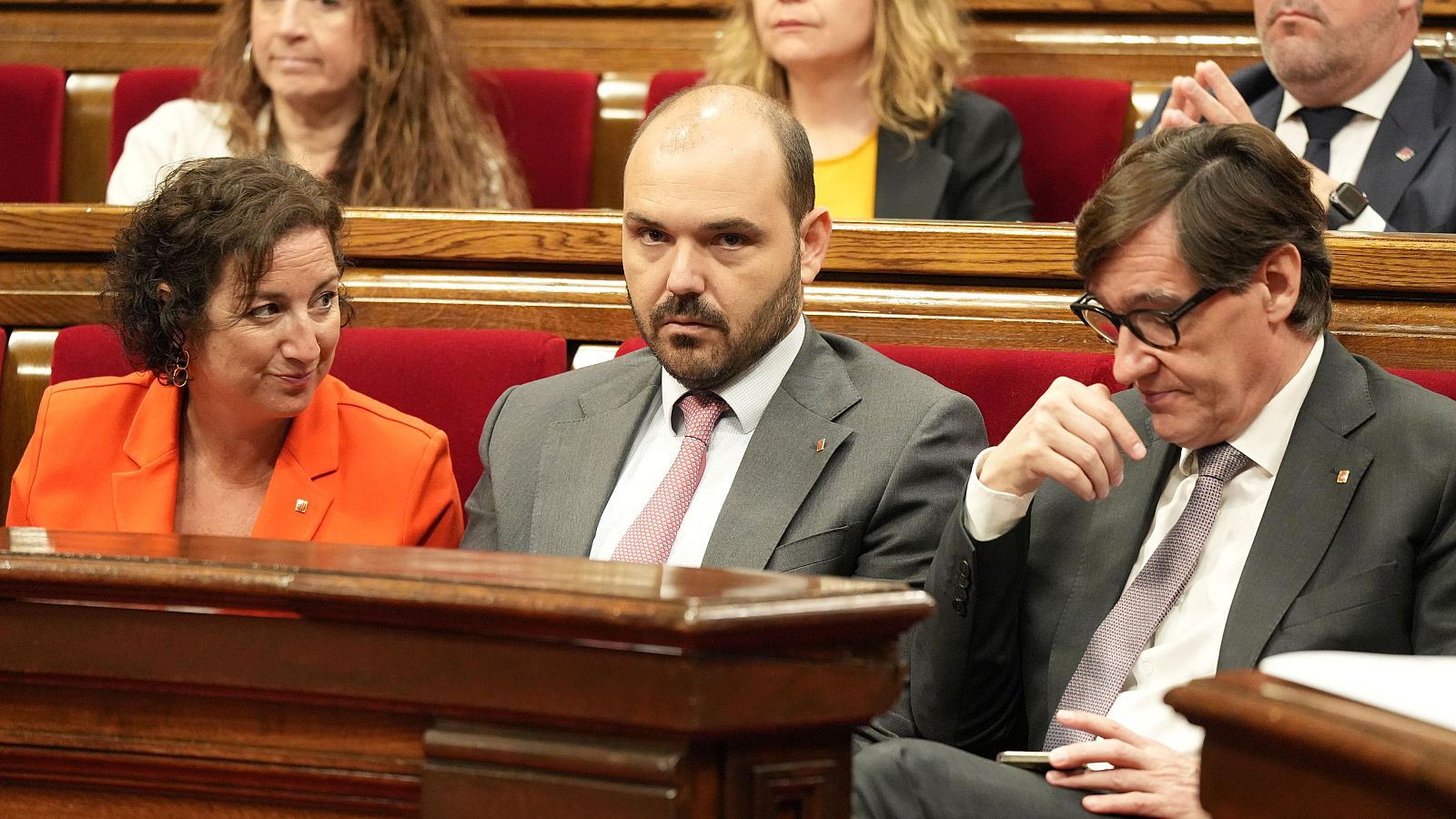 Alícia Romero, Albert Dalmau y Salvador Illa en un evento político. Romero (naranja), Dalmau (gris) y Illa (traje oscuro, gafas, móvil).