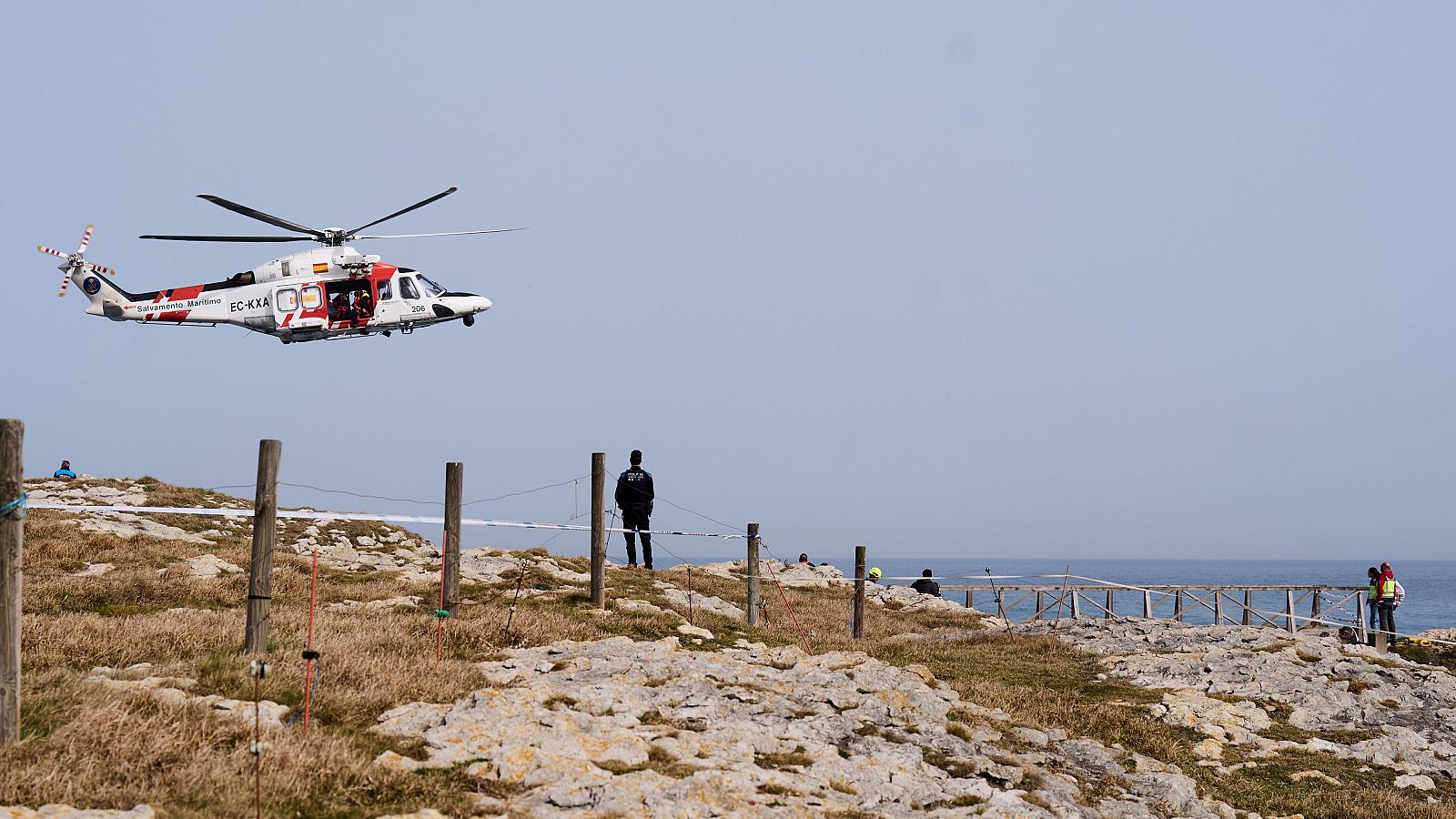 Helicóptero EC-KXA de Salvamento Marítimo en operación de rescate sobre zona costera rocosa. Personal de emergencia en tierra coordina la búsqueda.