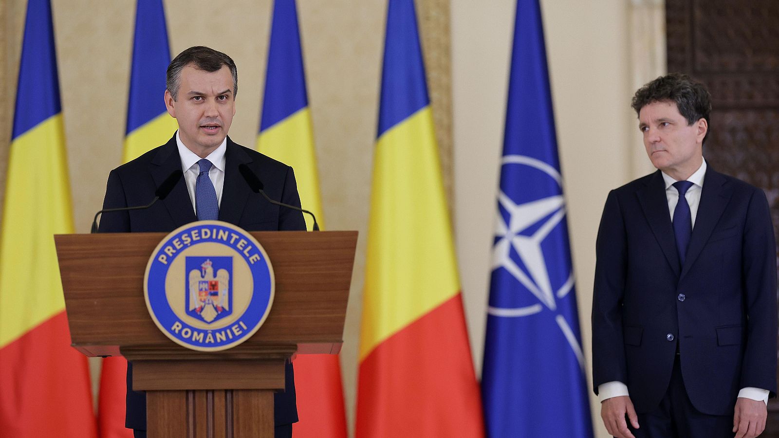 El presidente Nicușor Dan habla en un atril con el emblema "PREȘEDINTELE ROMÂNIEI", flanqueado por el nuevo primer ministro Eugen Tomac, con banderas de Rumanía y la OTAN al fondo.