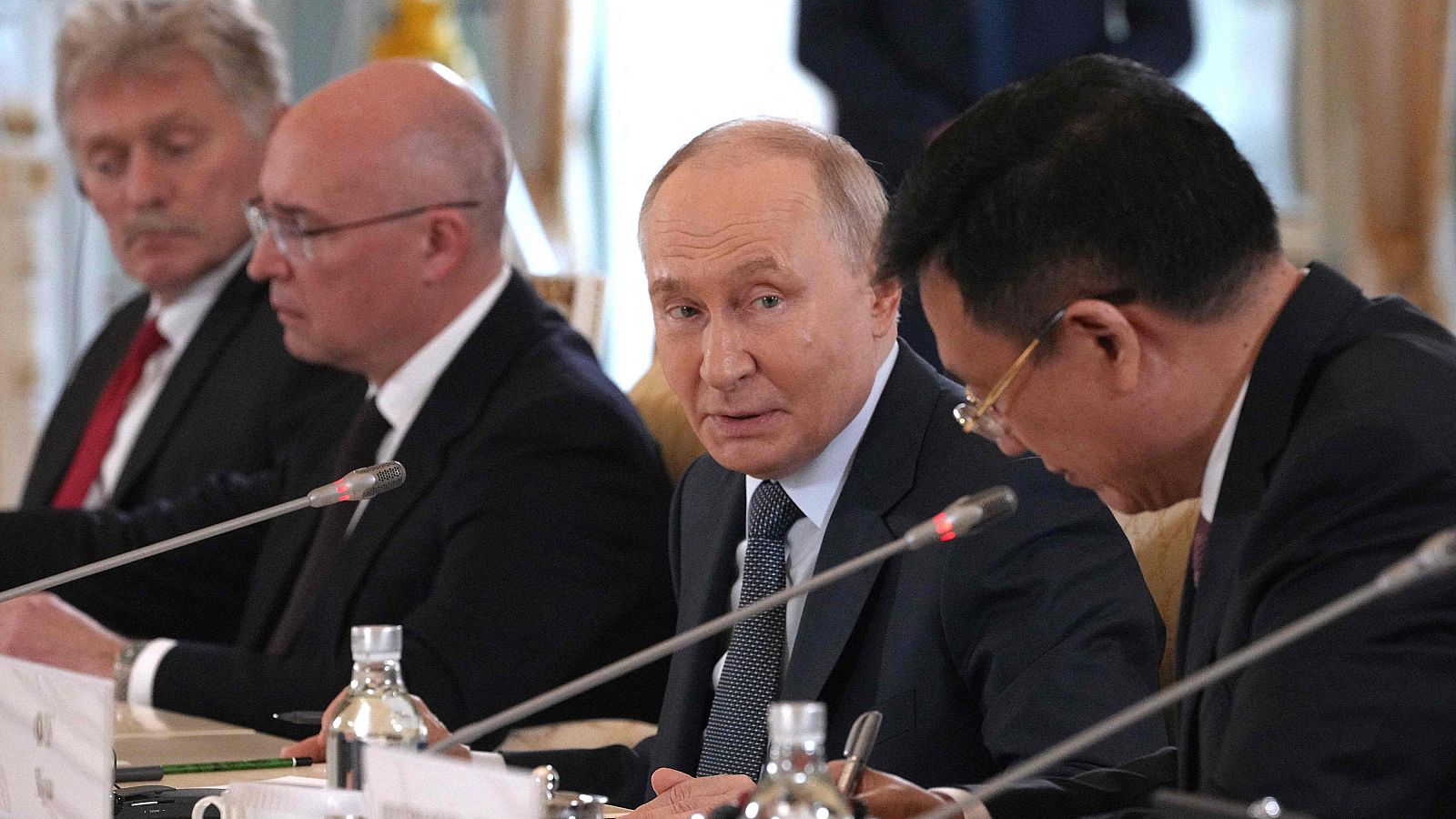 Vladimir Putin, presidente de Rusia, se reúne con Fu Hua, presidente de la Agencia de Noticias Xinhua, y otros representantes en una sala oficial. Se aprecian micrófonos y botellas de agua sobre la mesa.