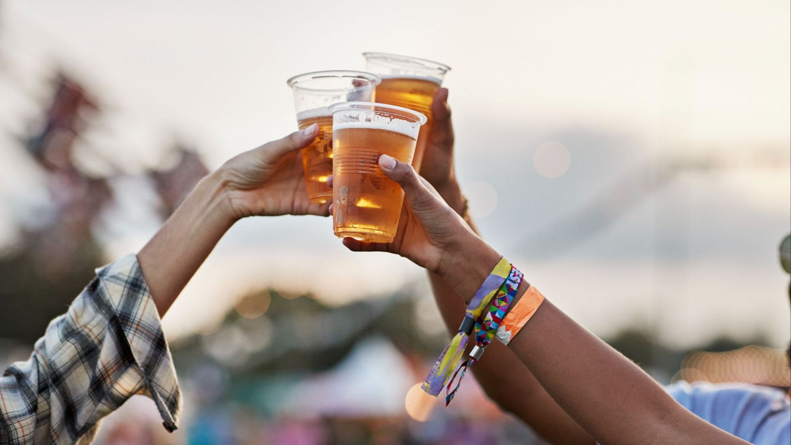Tres personas brindan con vasos de cerveza en un ambiente festivo. Se aprecian las manos sosteniendo los vasos en alto, con detalles como una camisa de cuadros y pulseras de colores.