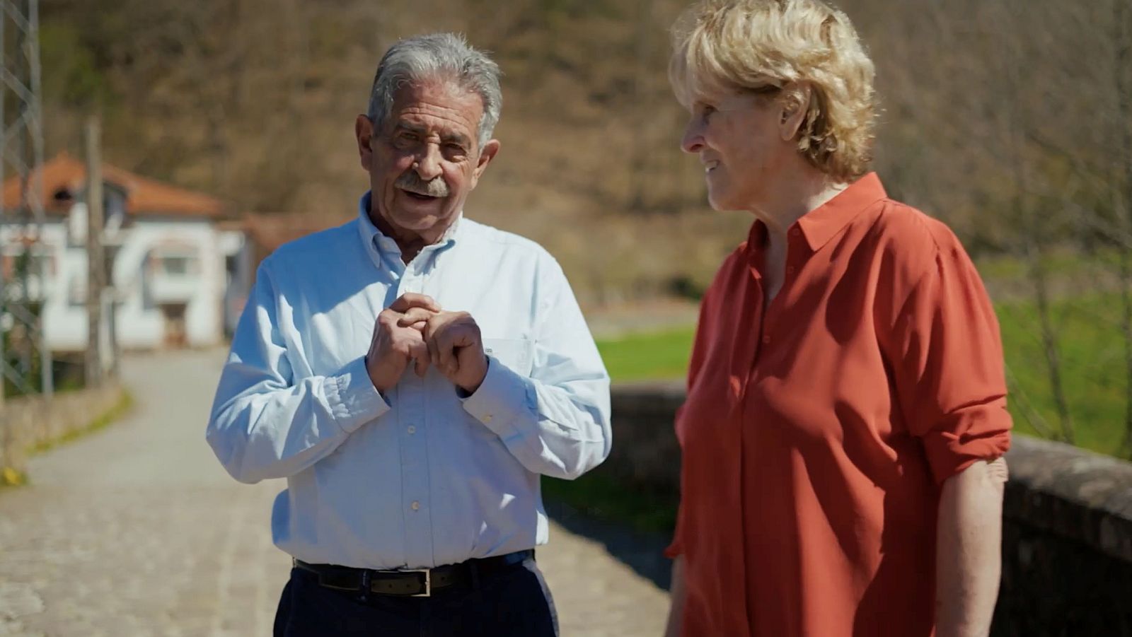 Miguel Ángel Revilla y Mercedes Milá conversan al aire libre, rodeados de un paisaje rural con edificaciones tradicionales. Revilla viste camisa azul y Milá una blusa naranja rojiza.