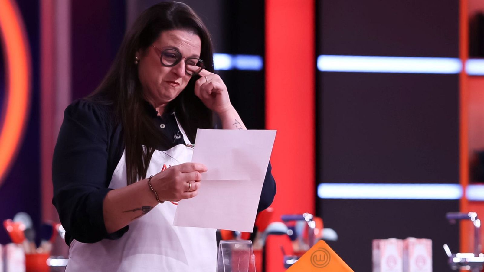 Lluís Puig, aspirante de MasterChef, llora de emoción mientras lee una carta en el plató del programa, rodeada de elementos icónicos del show.