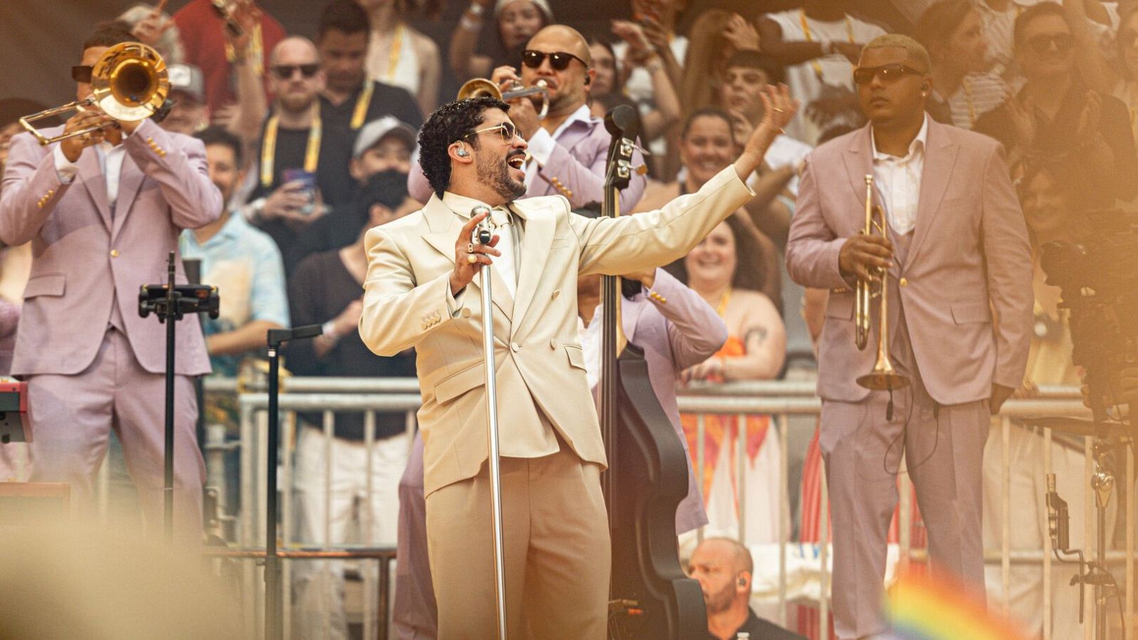 El cantante Bad Bunny, con traje de chaqueta color crema y gafas de sol, canta durante un concierto en el Riyadh Air Metropolitano, rodeado de músicos con trompetas y trajes rosa pálido.