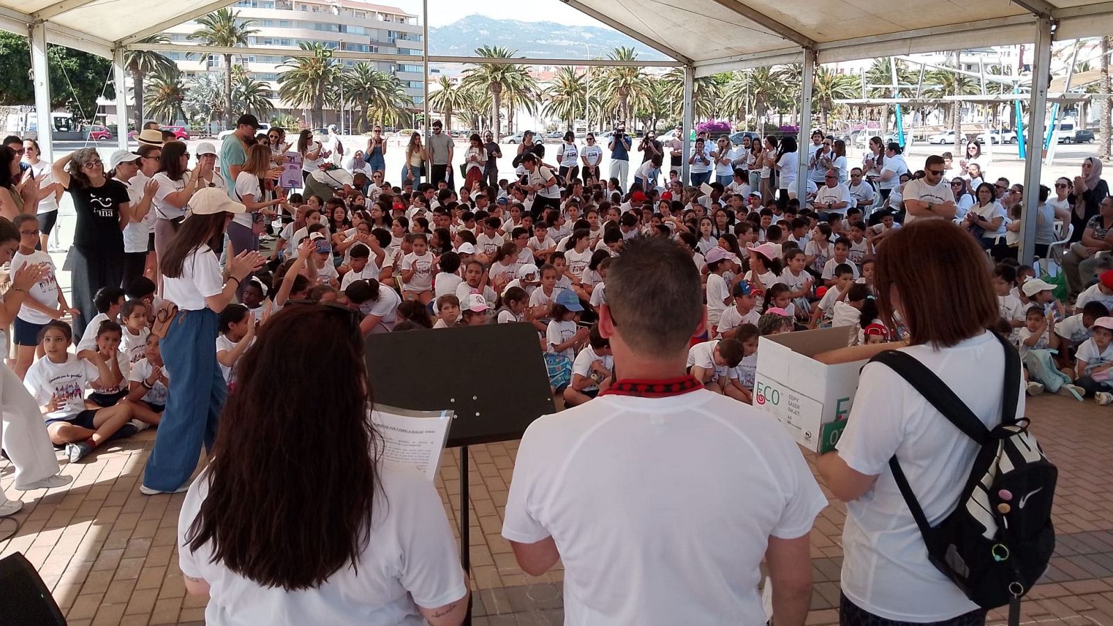 Evento al aire libre bajo carpa blanca con niños y adultos. Se observa una caja con "ECO" y palmeras al fondo. Fecha: 5 de junio de 2026, Melilla.