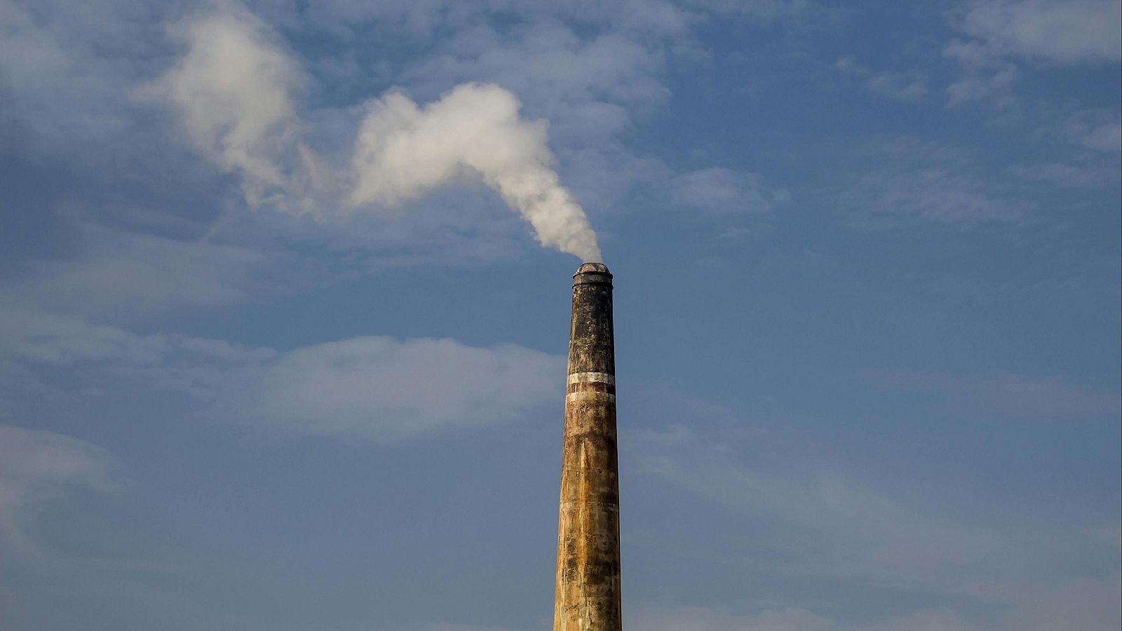 Imponente chimenea industrial de ladrillo con humo blanco ascendiendo hacia un cielo azul claro. Se aprecian bandas horizontales cerca de la parte superior.