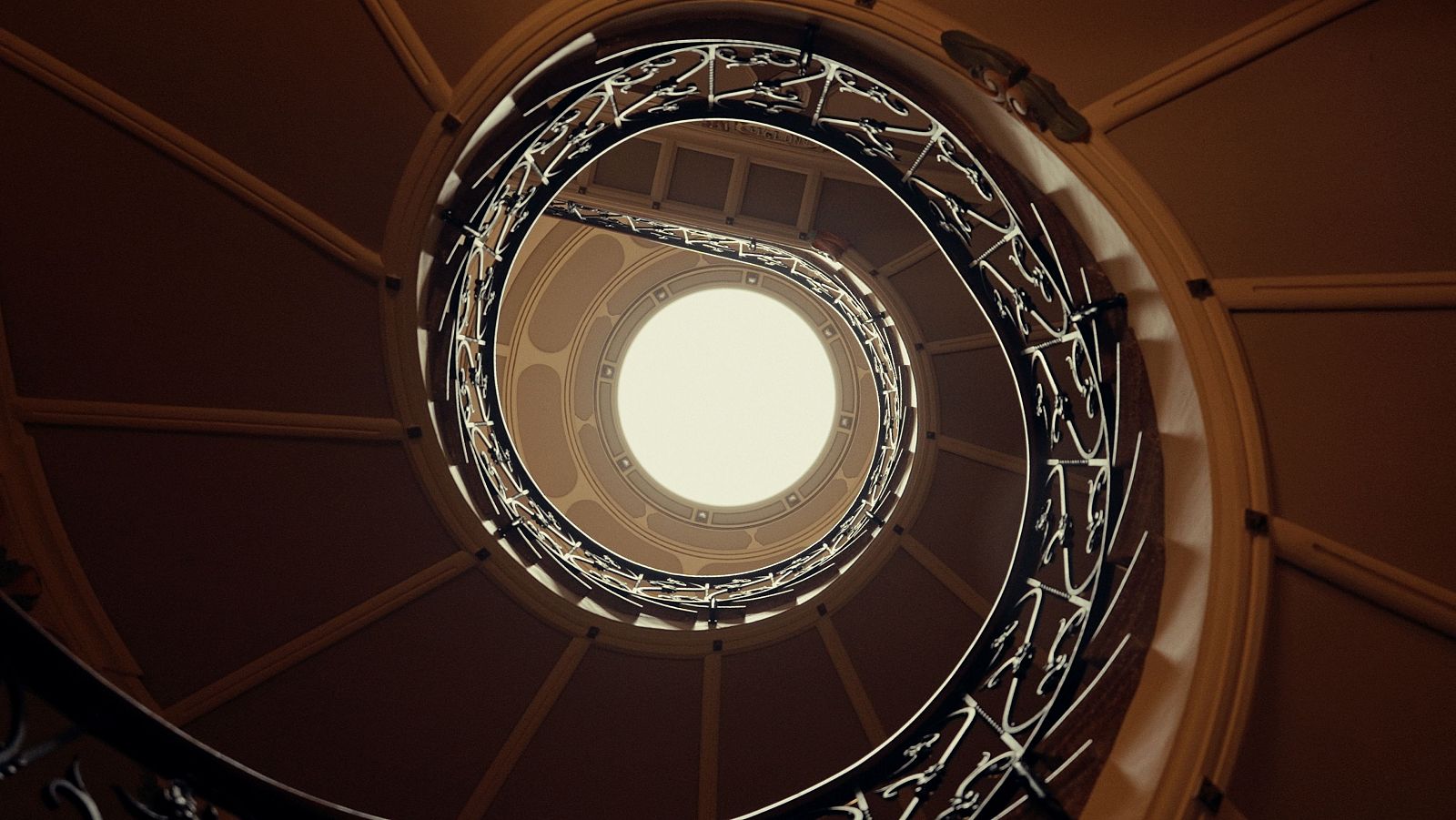 Escalera de caracol modernista con barandilla de hierro forjado ornamentada, ascendiendo hacia una luz cenital en el interior de Can Prunera, Sóller.