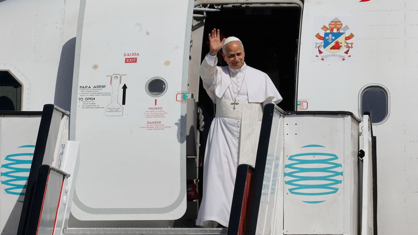 Papa Francisco saluda desde la puerta de un avión de ITA Airways, con escudo papal y advertencias de seguridad visibles.
