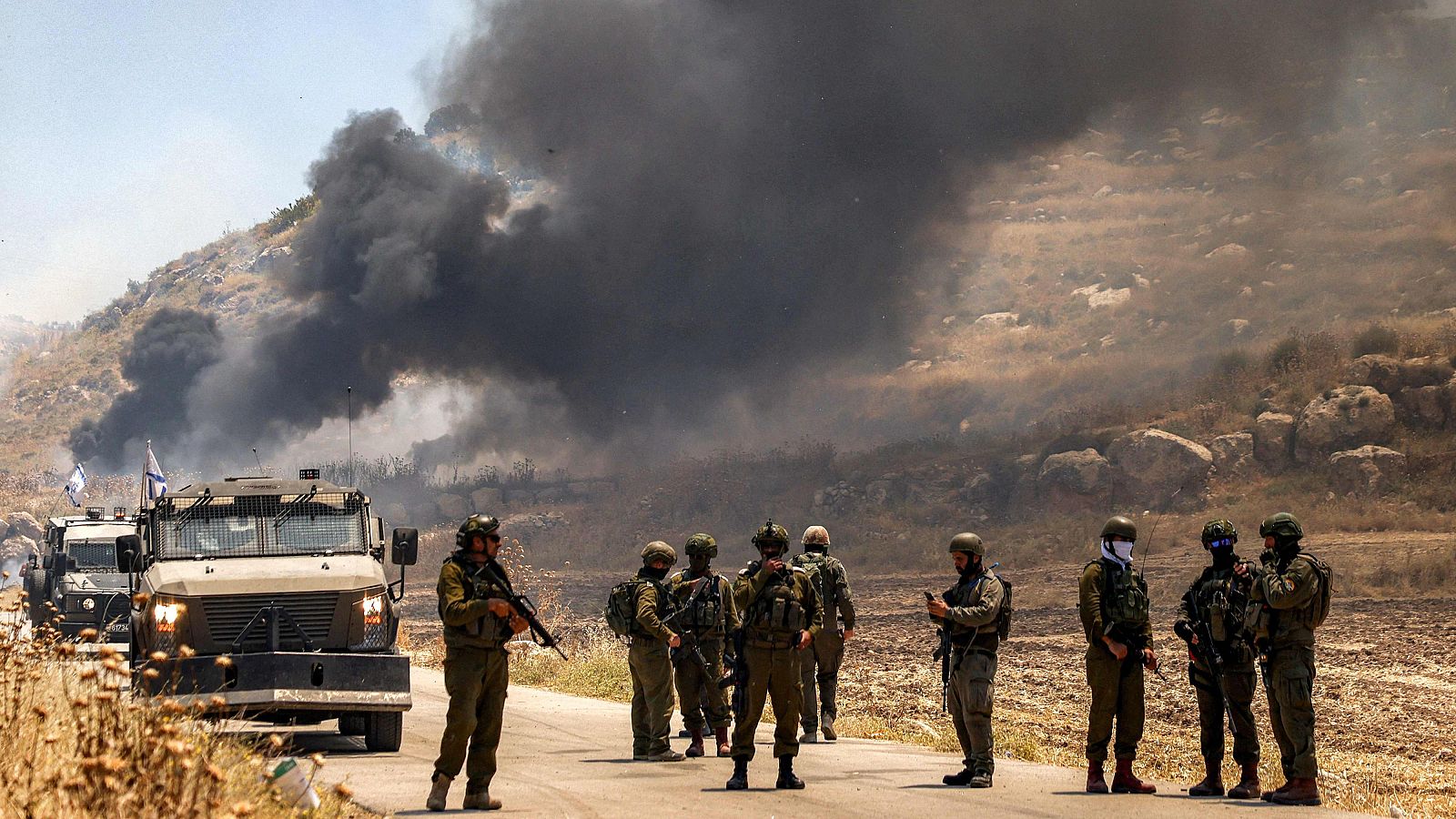 Fuerzas de seguridad israelíes hacen guardia junto a una carretera mientras, al fondo, se eleva humo procedente de una zona  incendiada por colonos israelíes en la aldea palestina de Idna, al oeste de Hebrón, en la Cisjordania ocupada.