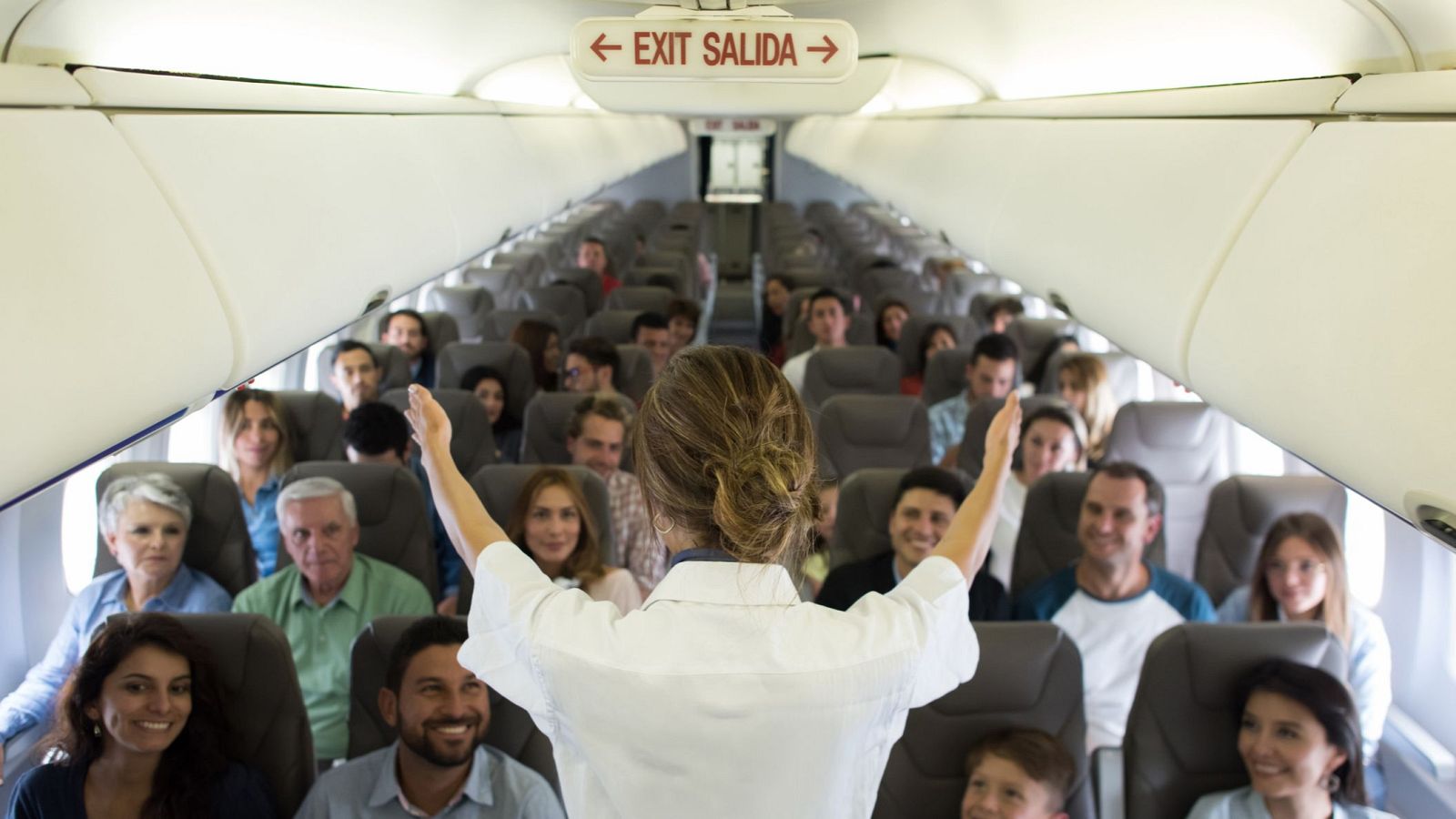 Interior de avión con tripulante de cabina dando instrucciones a pasajeros sonrientes. Se ve cartel "← EXIT SALIDA →".