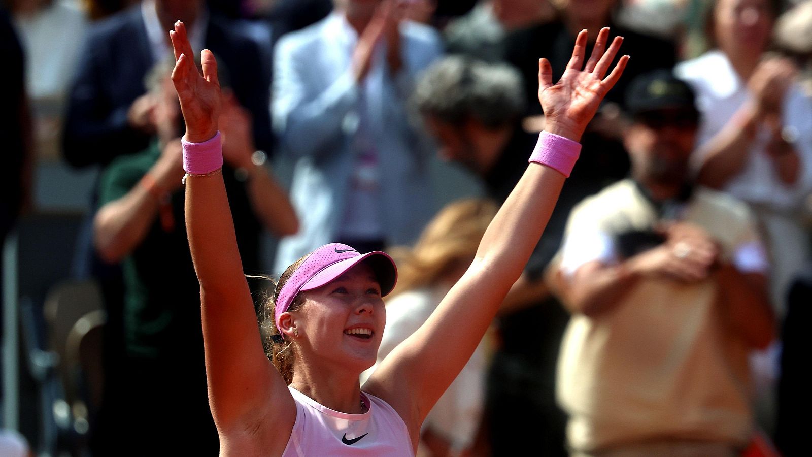 Mirra Andreeva, tenista, celebra su victoria en Roland Garros con los brazos en alto y una gran sonrisa. Lleva visera y camiseta rosa y blanca de Nike, y muñequeras rosas.