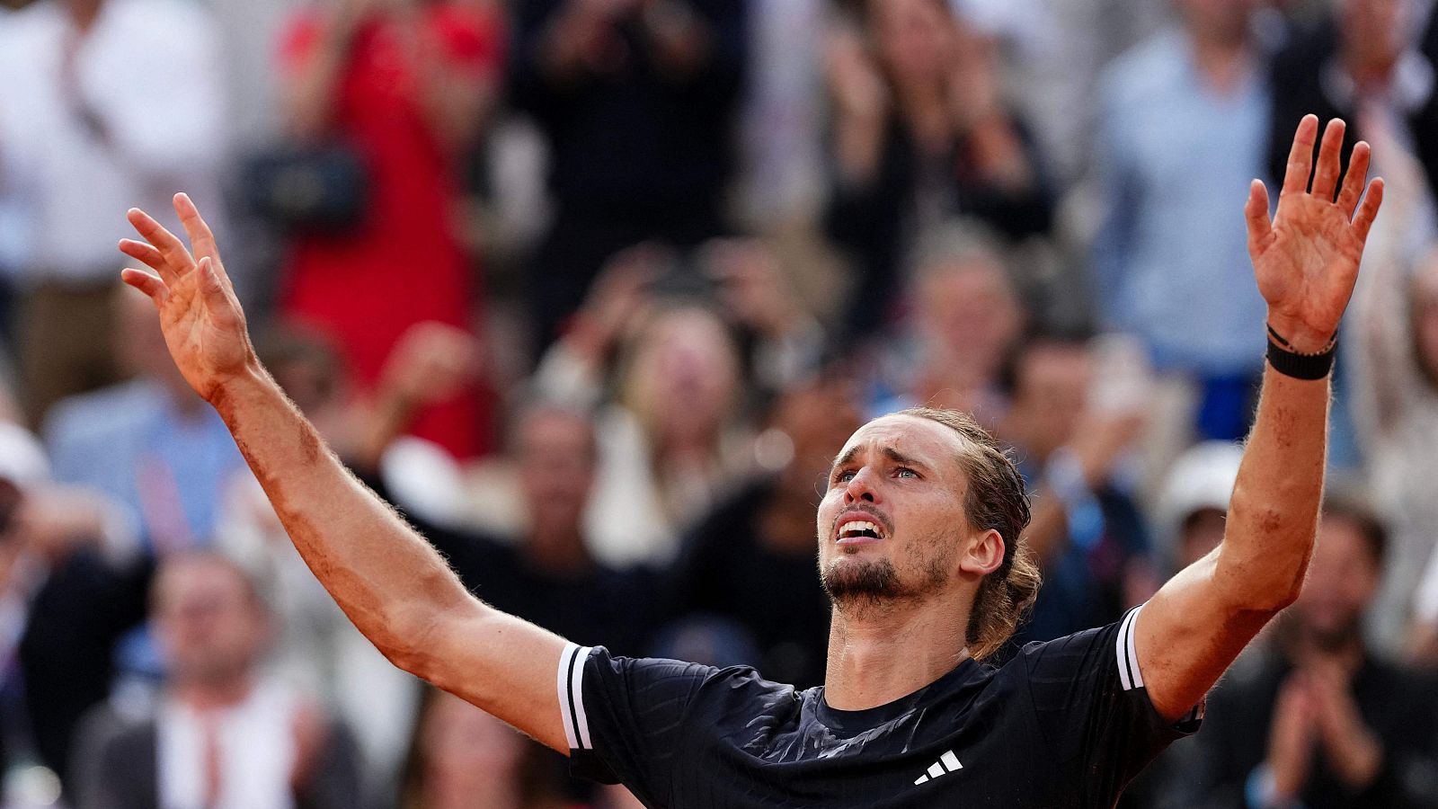 Alexander Zverev celebra su victoria en Roland Garros, con los brazos en alto y una expresión de éxtasis. Lleva una camiseta negra de Adidas.