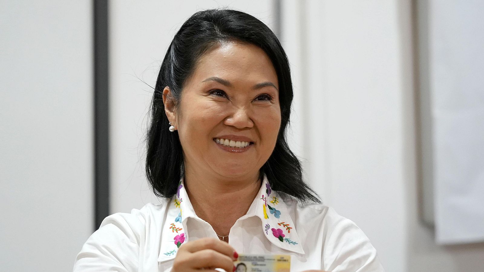 Keiko Fujimori sonríe y muestra su documento de identidad, vestida con una camisa blanca bordada y pendientes de perlas.