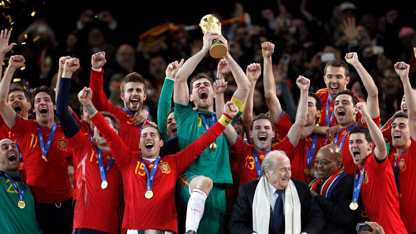 Iker Casillas levanta el trofeo de la Copa del Mundo rodeado de jugadores españoles eufóricos. Sepp Blatter y Jacob Zuma observan la celebración. Confeti dorado cae sobre la escena.