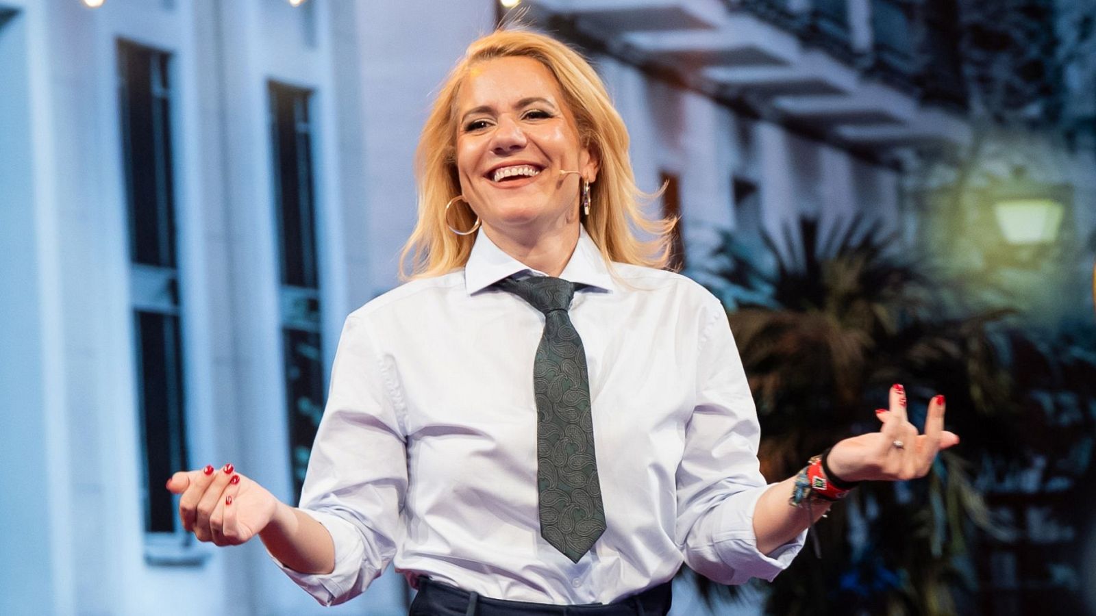 Henar Álvarez, rubia y sonriente, luce pendientes dorados, anillo y pulsera roja. Viste camisa clara y corbata gris oscuro, gesticulando con las manos.