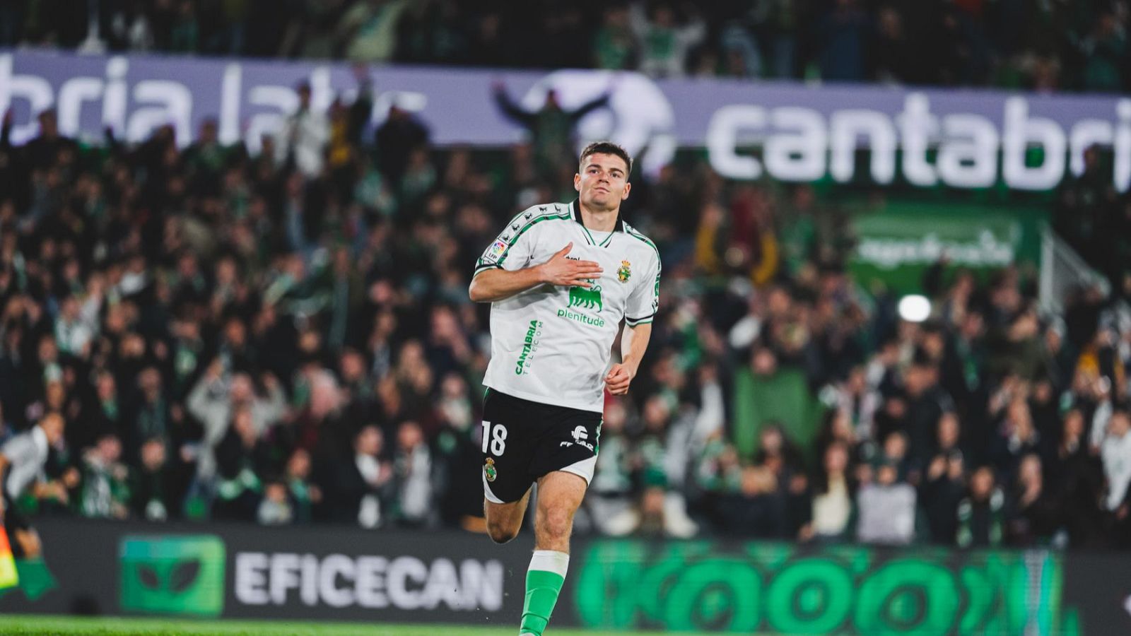 Peio Canales, jugador del Racing con el dorsal 18, celebra en el campo. Viste camiseta blanca con "Plenitude" y "CANTABRIA", rodeado de aficionados y vallas publicitarias.