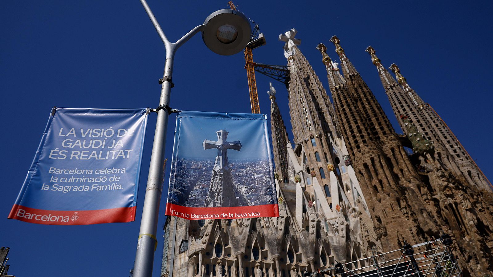 Vista de la Sagrada Familia con pancartas informativas y una grúa de construcción, bajo un cielo azul. Destaca el texto 'LA VISIÓ DE GAUDÍ JA ÉS REALITAT' y 'Fem la Barcelona de la teva vida.'