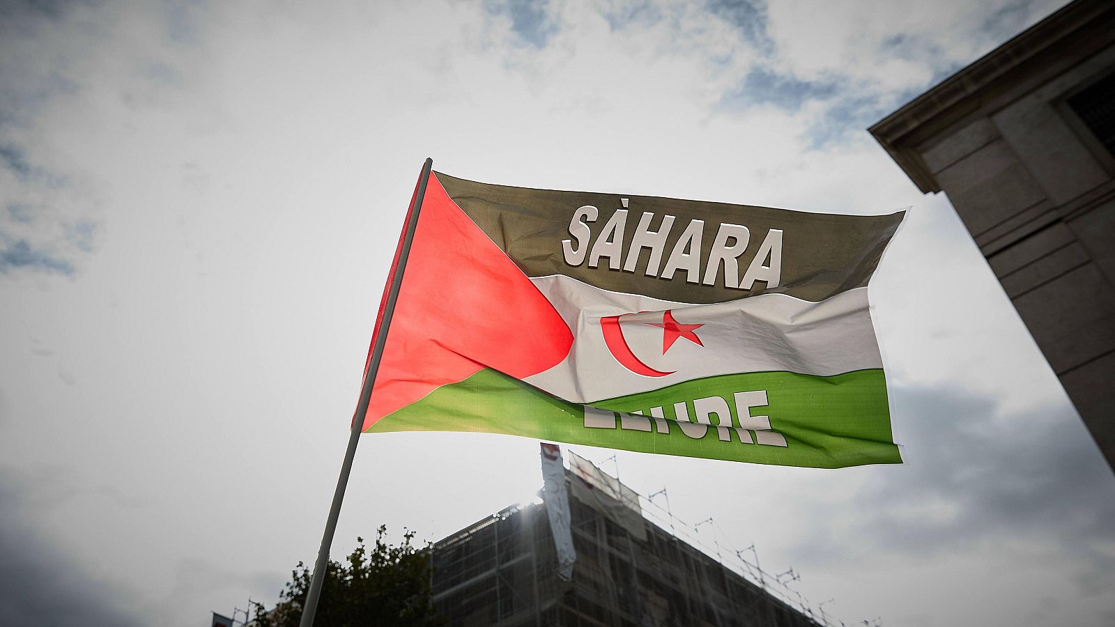 Bandera del Sáhara Occidental con "SÁHARA LIBRE" en blanco, triángulo rojo, franjas de colores, cielo nublado y siluetas urbanas.