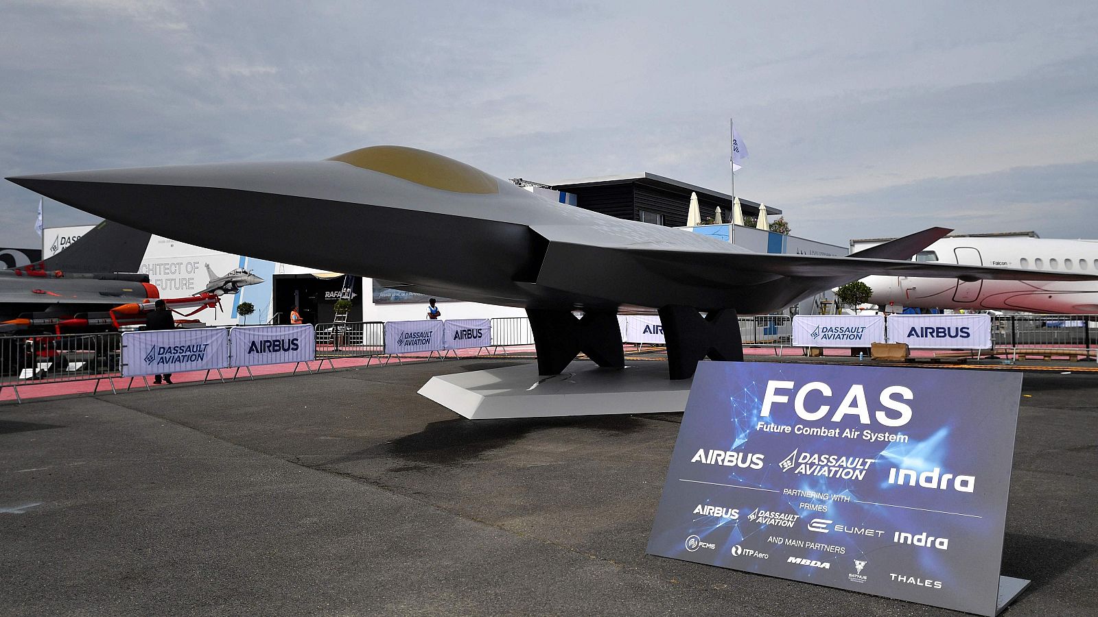 Maqueta a escala del avión de combate furtivo FCAS, expuesta al aire libre con cartel informativo de AIRBUS, DASSAULT AVIATION e indra. Entorno de feria aeroespacial con vallas y otras maquetas.