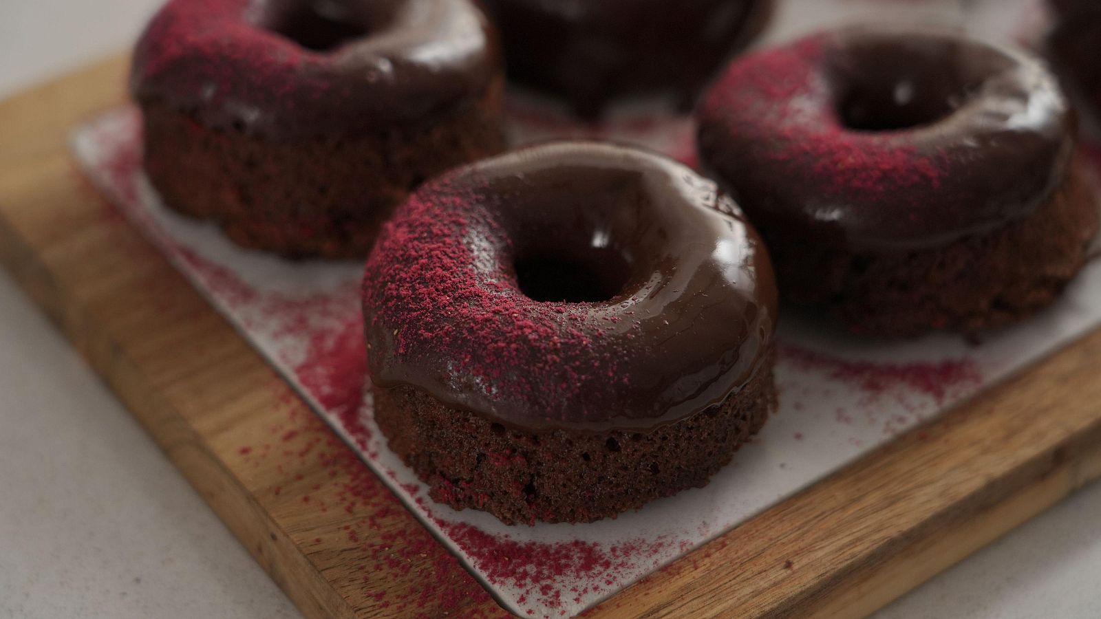 Primer plano de donuts de chocolate con glaseado brillante y espolvoreado rosa/rojo sobre papel de horno y tabla de madera.