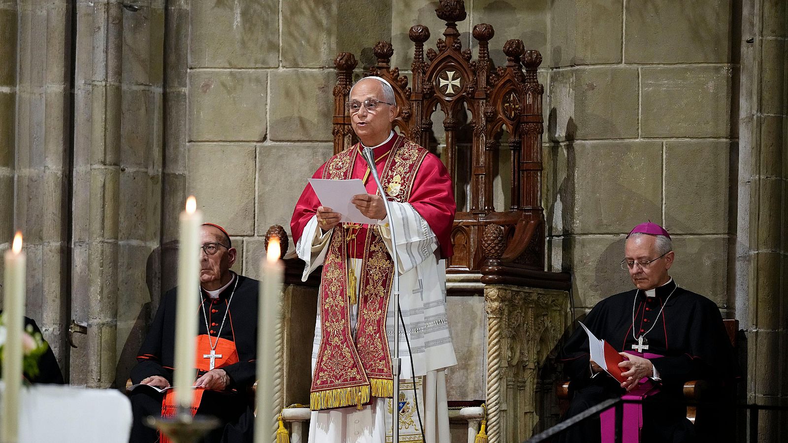 El Papa León XIV oficia un rezo en la Catedral de Barcelona, acompañado por un cardenal y un obispo. La escena se desarrolla en un ambiente solemne con arquitectura gótica y velas encendidas.