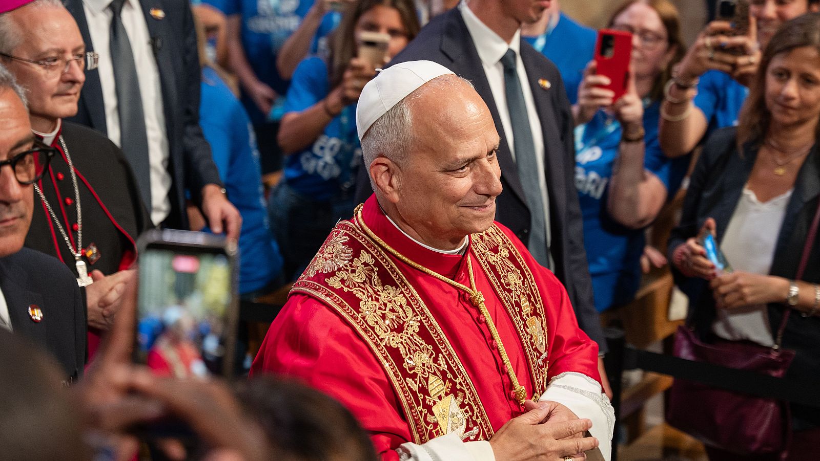 Primer plano del Papa Lleó XIV con casulla roja, junto a un clérigo de edad avanzada. Multitud al fondo con móviles y camisas azules.