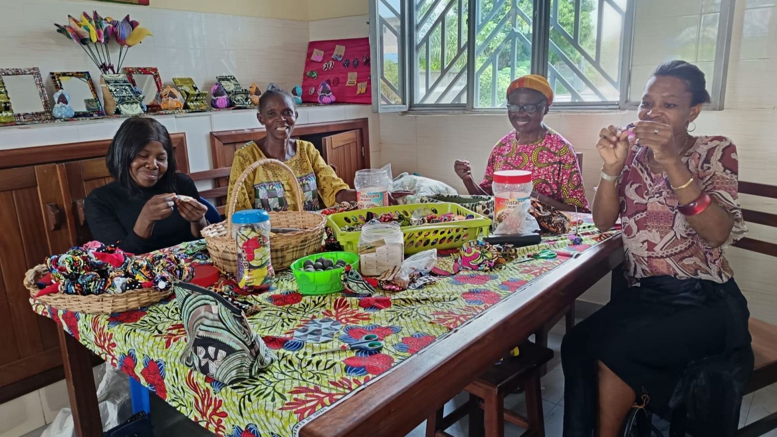 Cuatro mujeres de piel oscura y una de piel más clara, sentadas alrededor de una mesa con mantel colorido, disfrutan de una actividad de manualidades con diversos materiales.