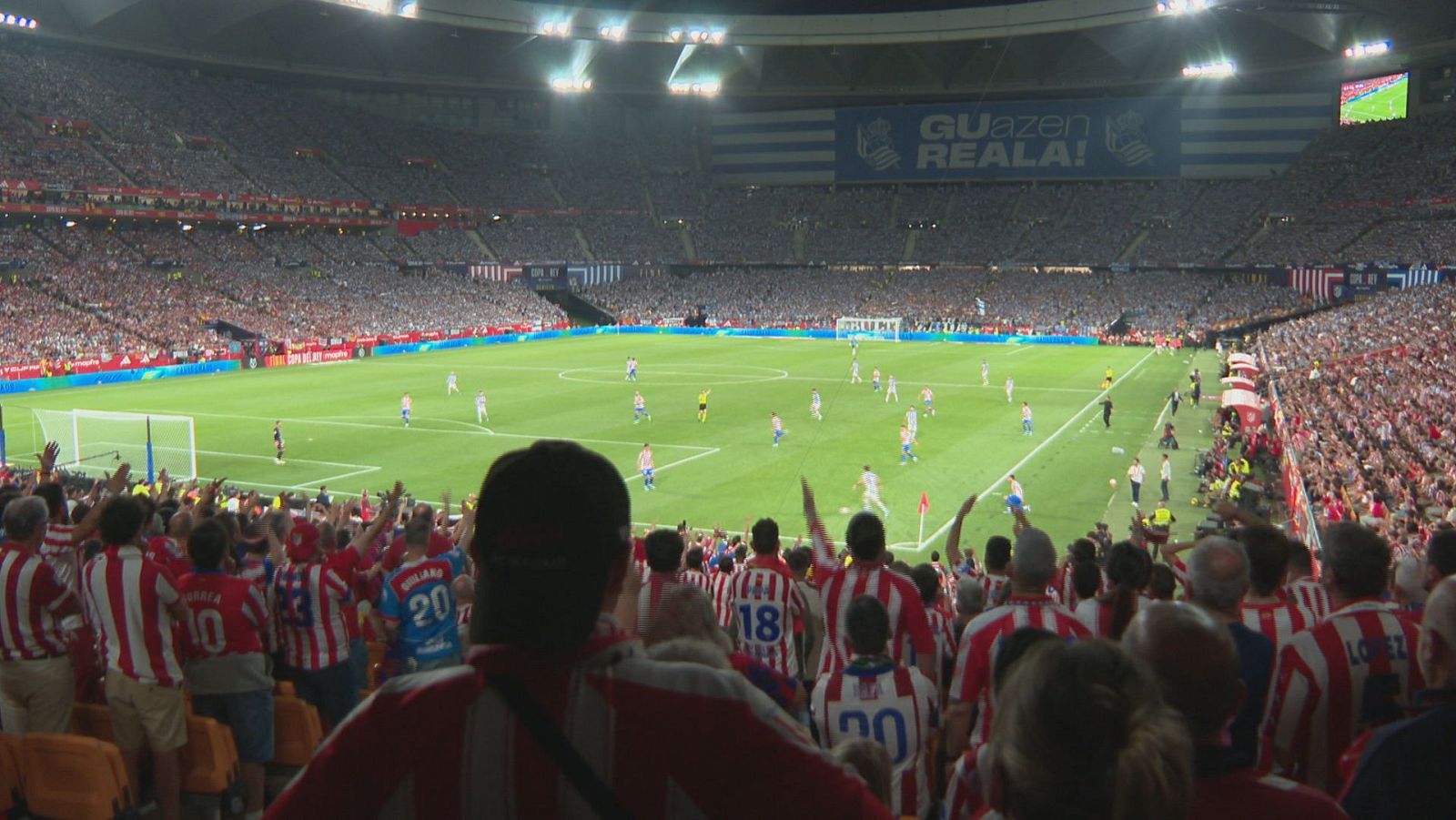 Vista aérea de un estadio de fútbol nocturno con miles de aficionados. Jugadores en el campo, uno con camiseta a rayas rojas, blancas y azules. Aficionados en gradas rojas y blancas animan, algunos con camisetas del '10', '18', '20'. Pancarta azul y blanca con 'GUAzen REALA!'.