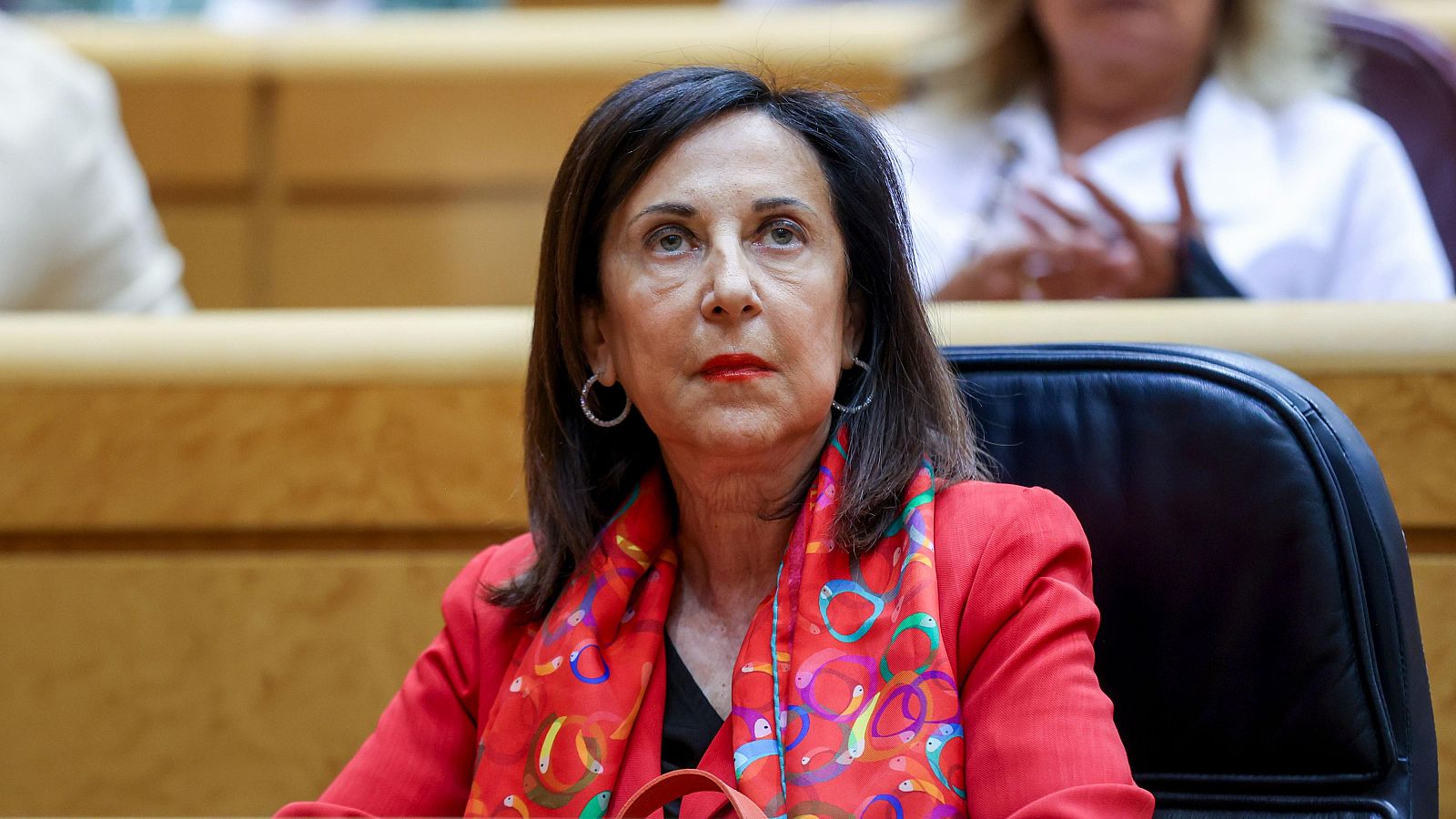 Primer plano de Margarita Robles, Ministra de Defensa de España, en el Senado. Viste chaqueta roja, bufanda colorida y pendientes plateados. Mira atentamente hacia arriba.