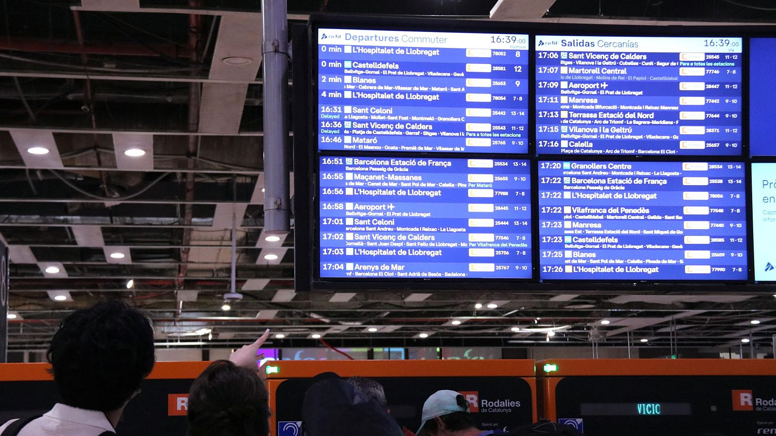 Dos personas observan pantallas azules con información de salidas de trenes de cercanías en una estación. Se ve la hora "16:39" y destinos como L'Hospitalet de Llobregat.