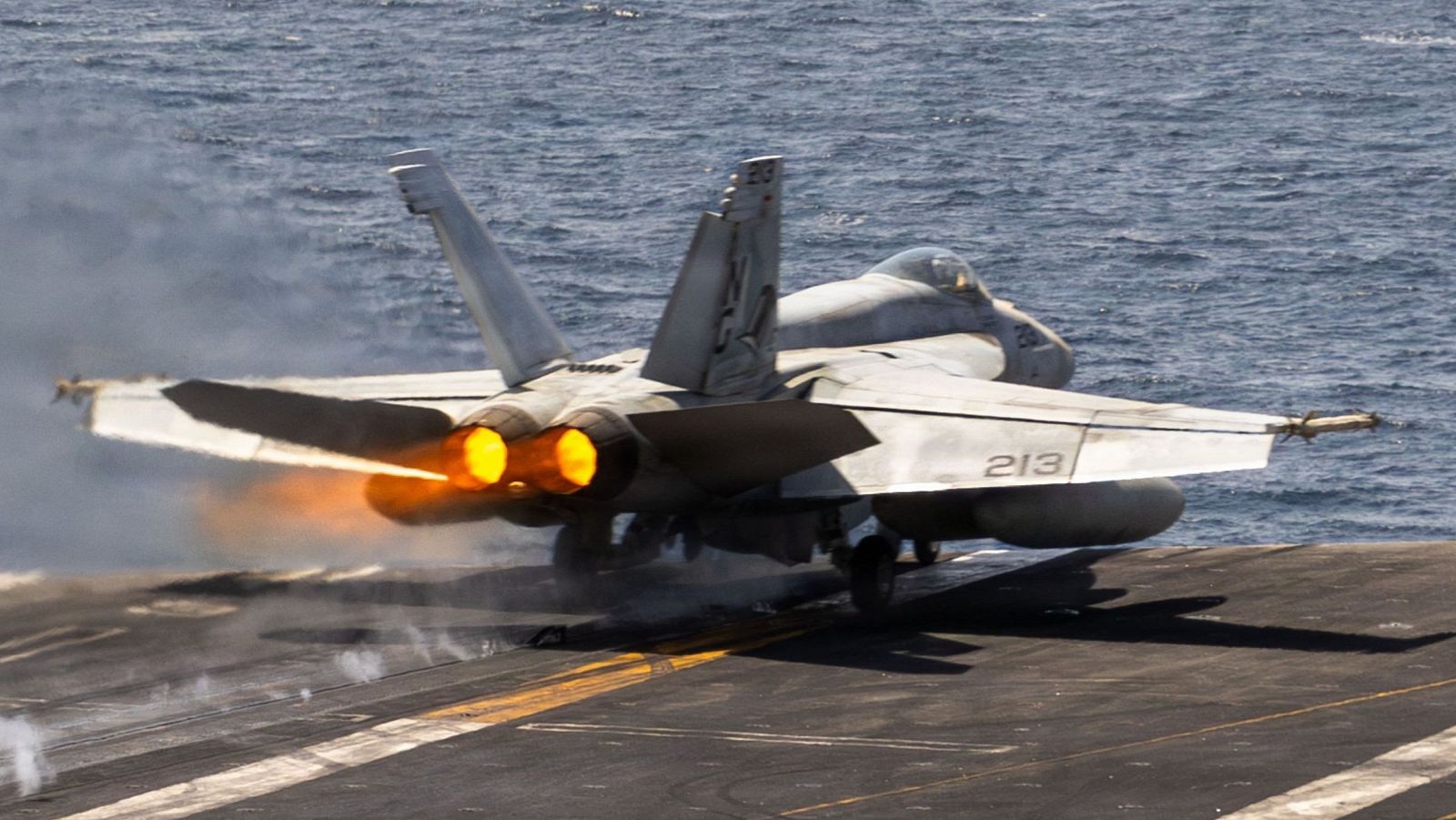 Avión F-18 con el número "213" en ala derecha, despegando con postquemadores activados. Humo y vapor en cubierta oscura de portaaviones sobre el mar.