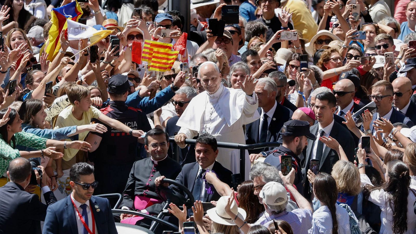El Papa Francisco saluda a una multitud con banderas de Cataluña y del Vaticano. Se observa personal de seguridad y un clérigo en un vehículo.