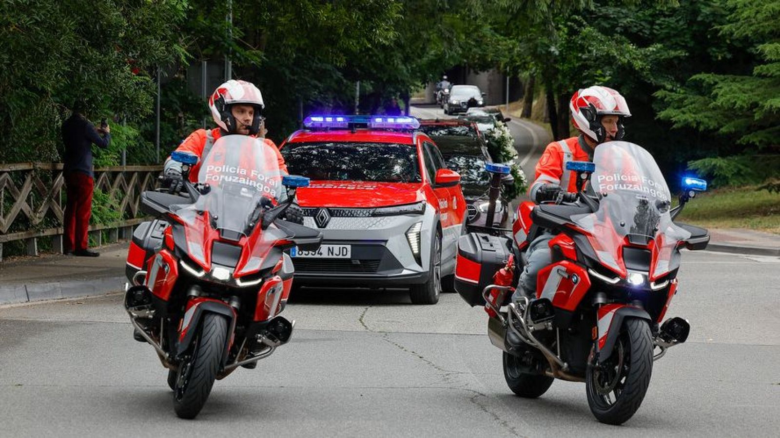 Motocicletas de la Policía Foral de Navarra escoltan un cortejo fúnebre en una carretera. Se aprecian detalles de los uniformes y vehículos policiales con luces azules.