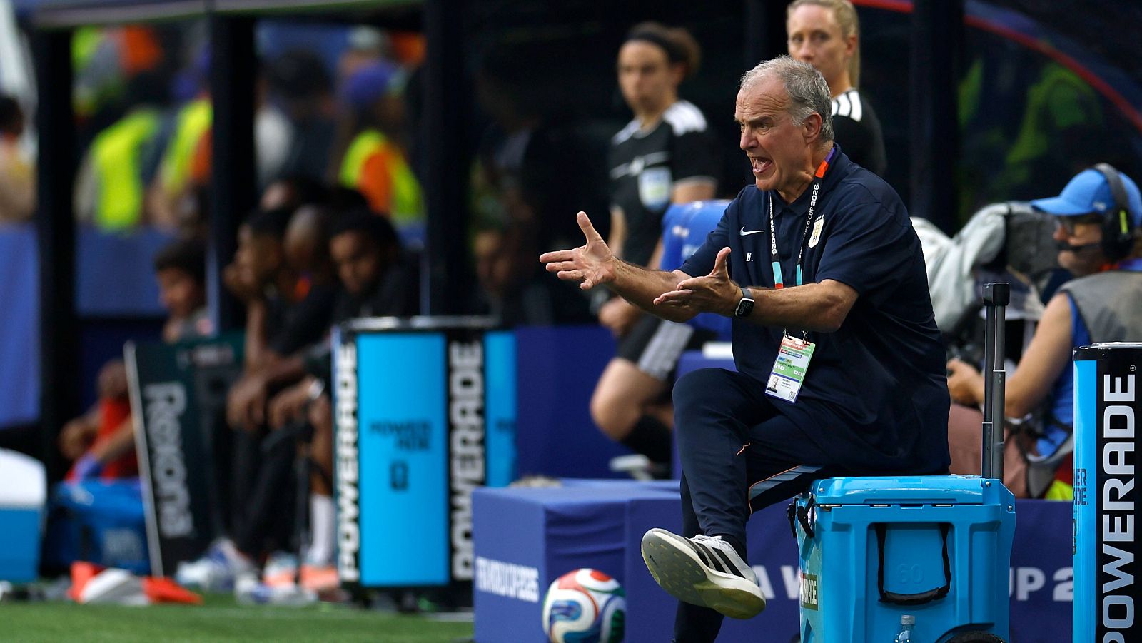 Marcelo Bielsa, seleccionador de Uruguay, sentado en un enfriador azul, gesticula con frustración en la línea de banda de un campo de fútbol.