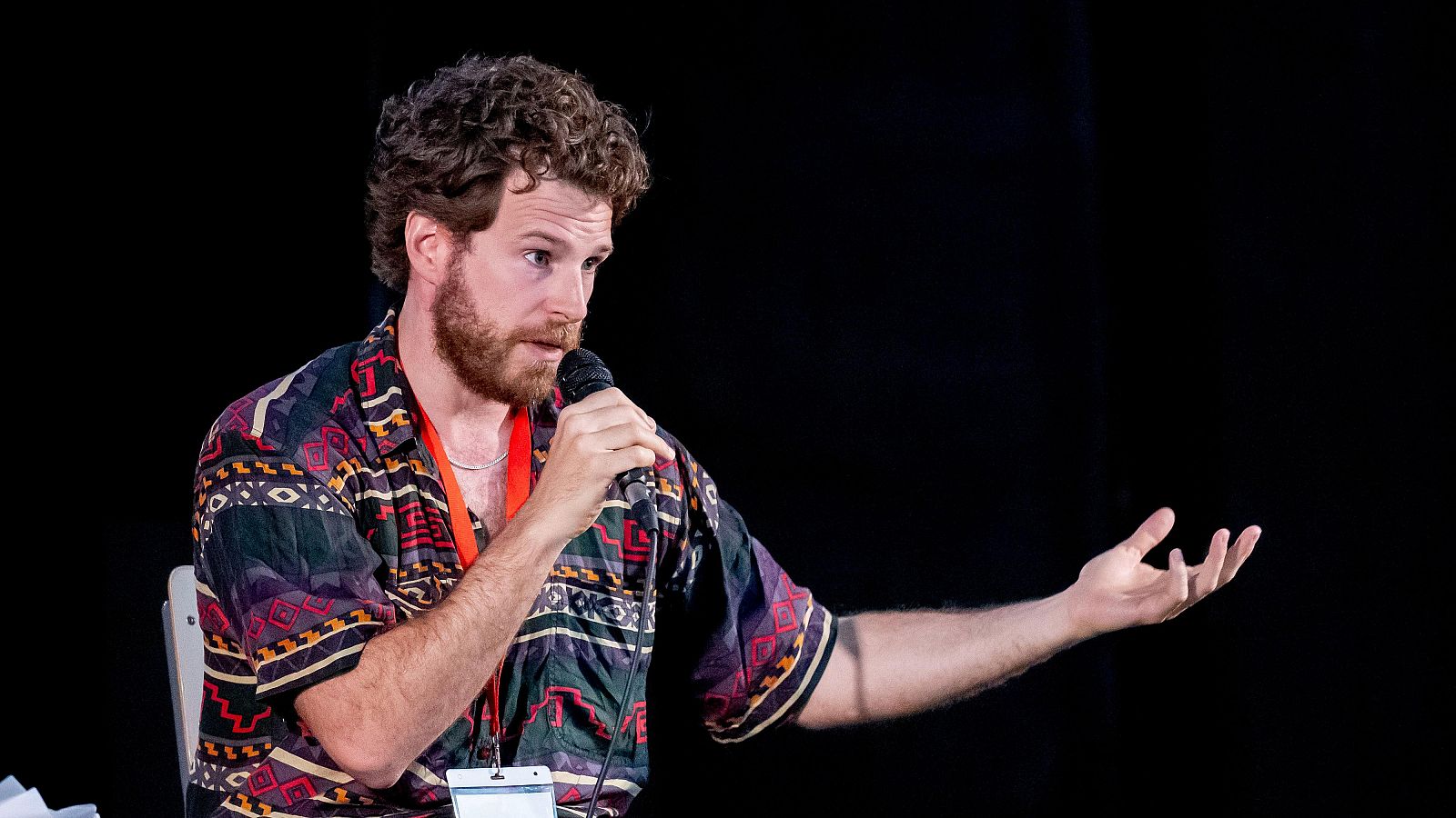 Álvaro Cervantes, actor, con cabello rizado y barba, habla en un coloquio. Sostiene un micrófono y gesticula con la mano izquierda. Viste camisa colorida y lleva una cinta naranja de acreditación.
