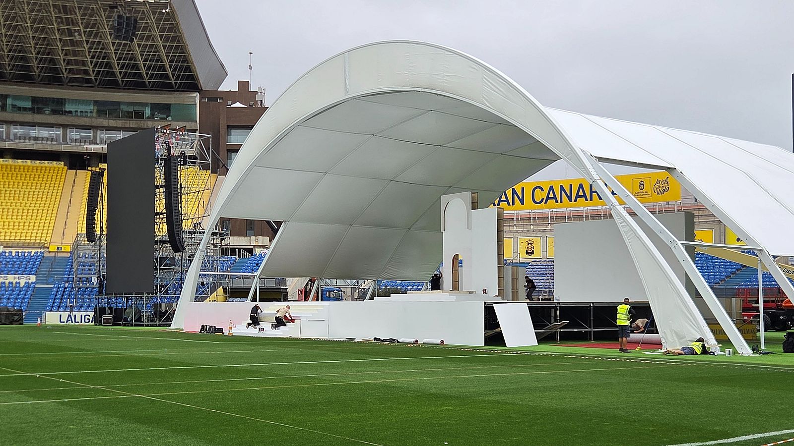 Vista panorámica de un estadio de fútbol con montaje de escenario, pantallas LED y equipo de sonido. Se aprecian las gradas con "GRAN CANARIA" y "LALIGA". Personas trabajan en los preparativos bajo un cielo nublado.