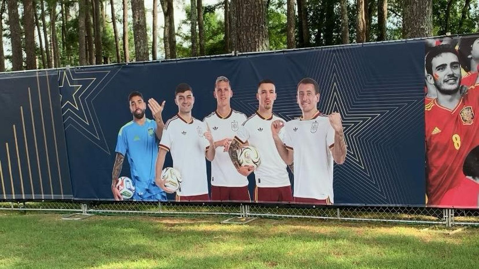 Lona publicitaria con futbolistas de la selección española: Pedri, Dani Olmo y Gavi. Fondo azul con estrellas doradas. Césped y árboles al fondo.