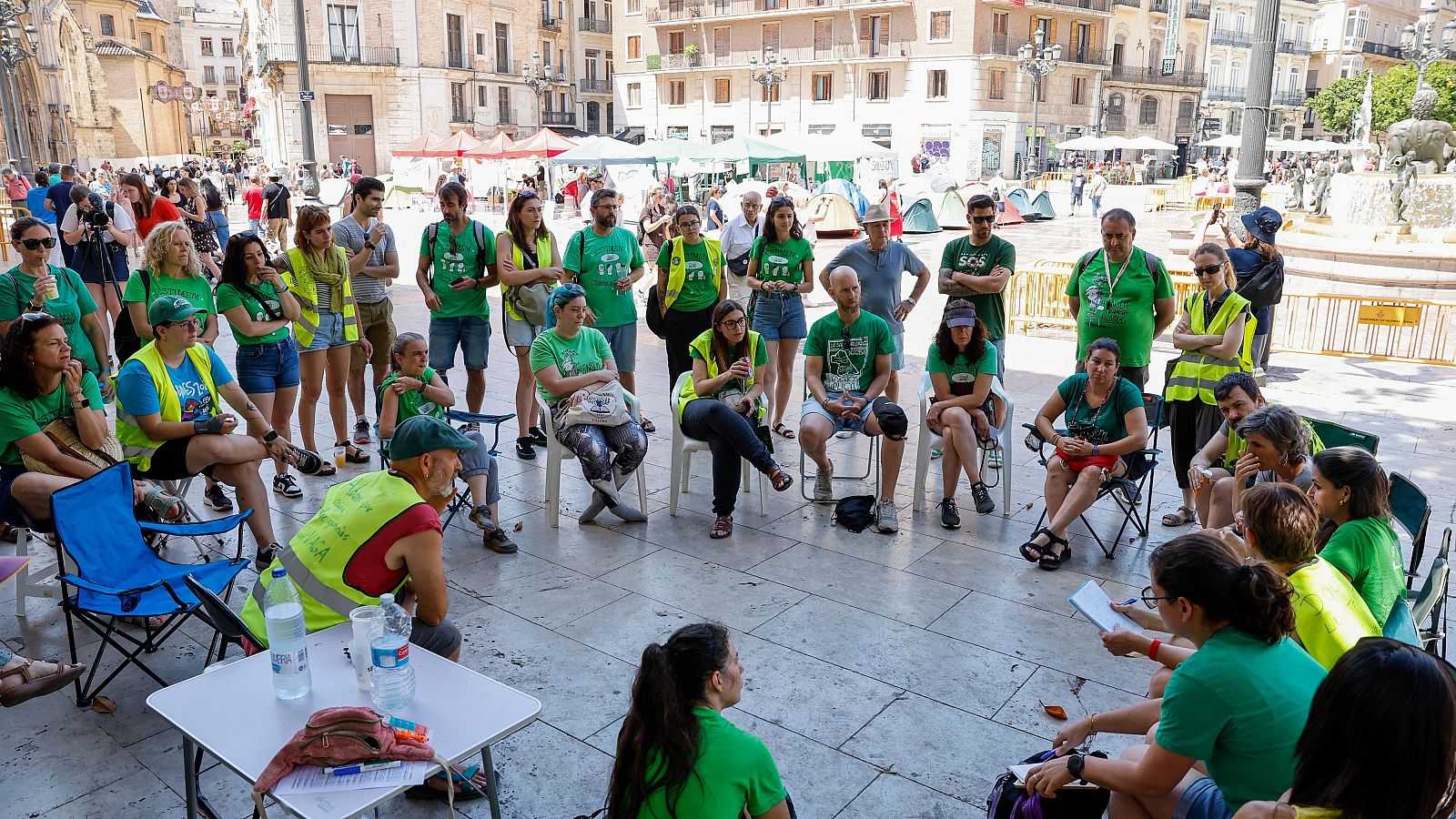 Asamblea de docentes en València con camisetas "SOS PÚBLICA" y chalecos reflectantes, rodeados de tiendas de campaña en una plaza céntrica.