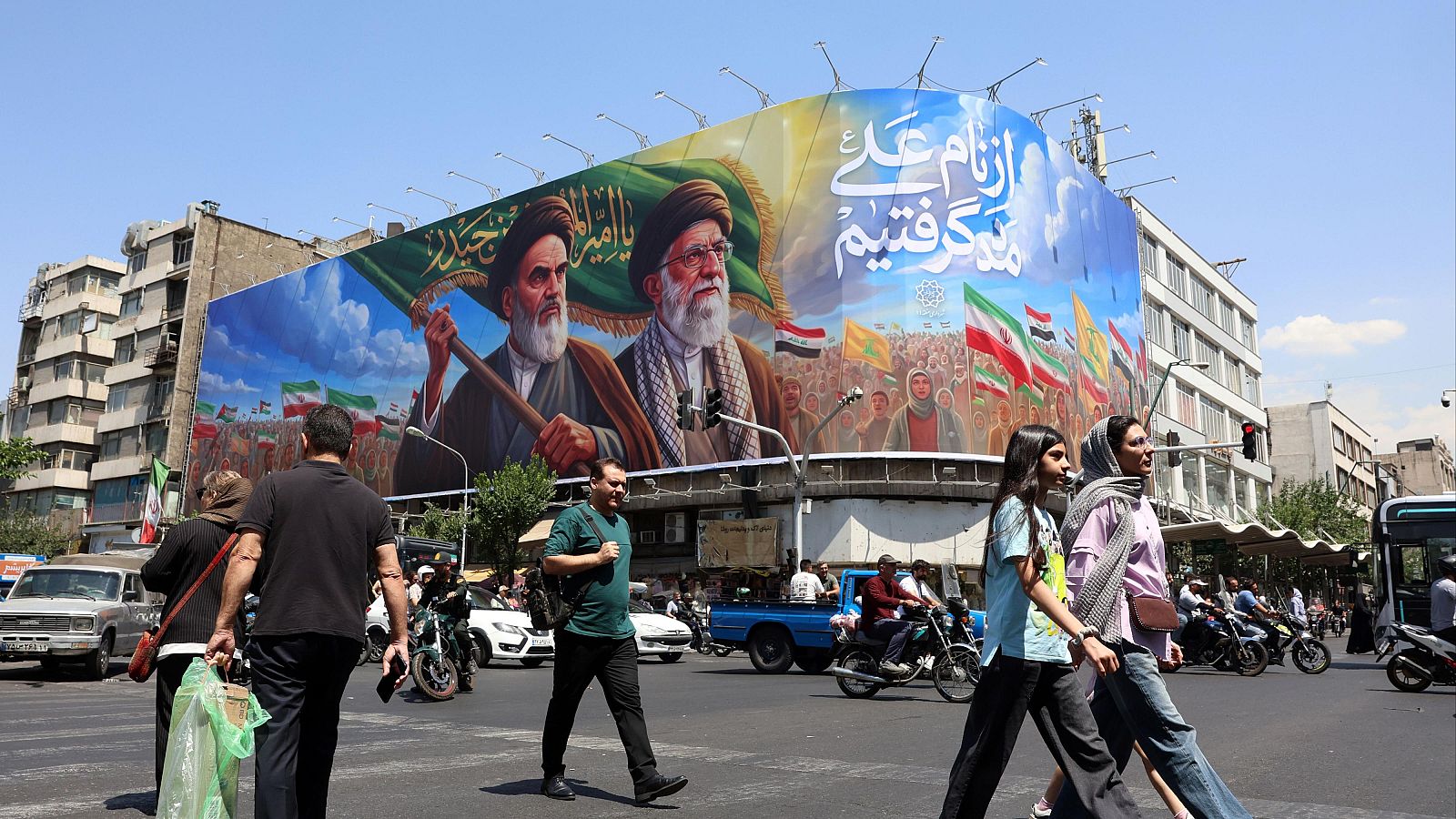 Valla publicitaria gigante en Teherán con Jomeini y Jamenei, rodeados de banderas iraníes y caligrafía árabe. Peatones y tráfico en la calle bajo un cielo azul.