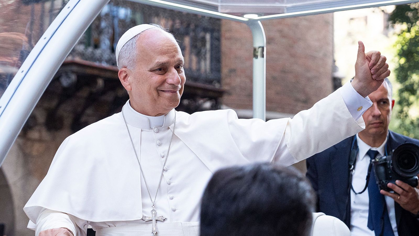 Hombre con atuendo papal blanco, sonríe y levanta el pulgar, dentro de un vehículo con techo transparente. Un fotógrafo con cámara profesional se ve detrás.