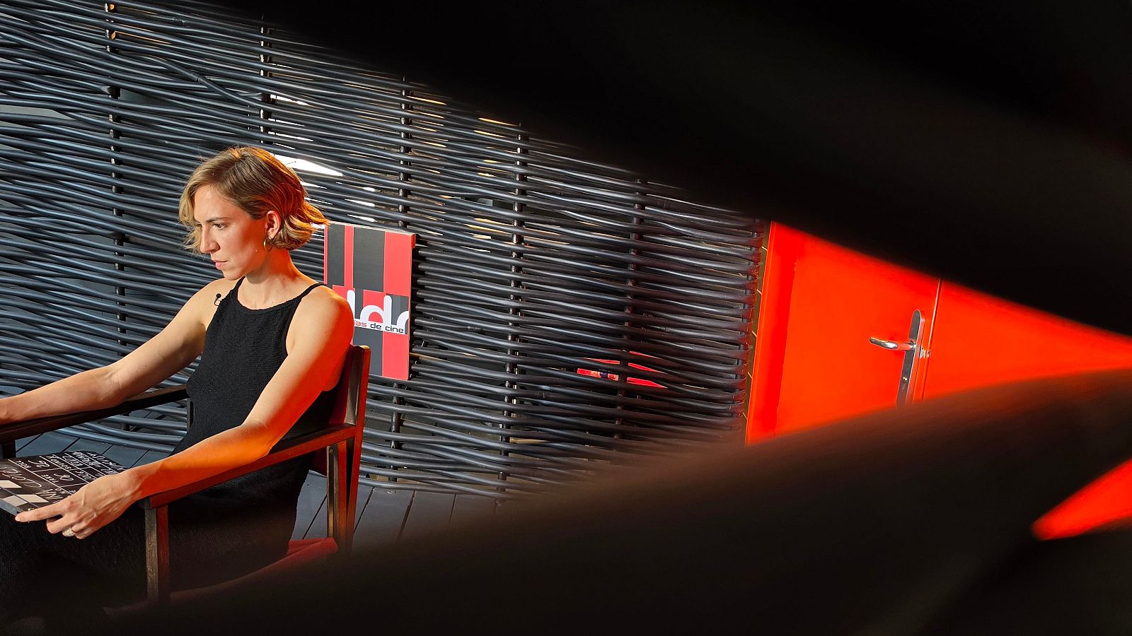 Aina Clotet, con top negro y pelo corto rubio ceniza, sentada en silla de madera oscura, mira hacia abajo con expresión concentrada. Sus manos sobre un objeto plano oscuro.