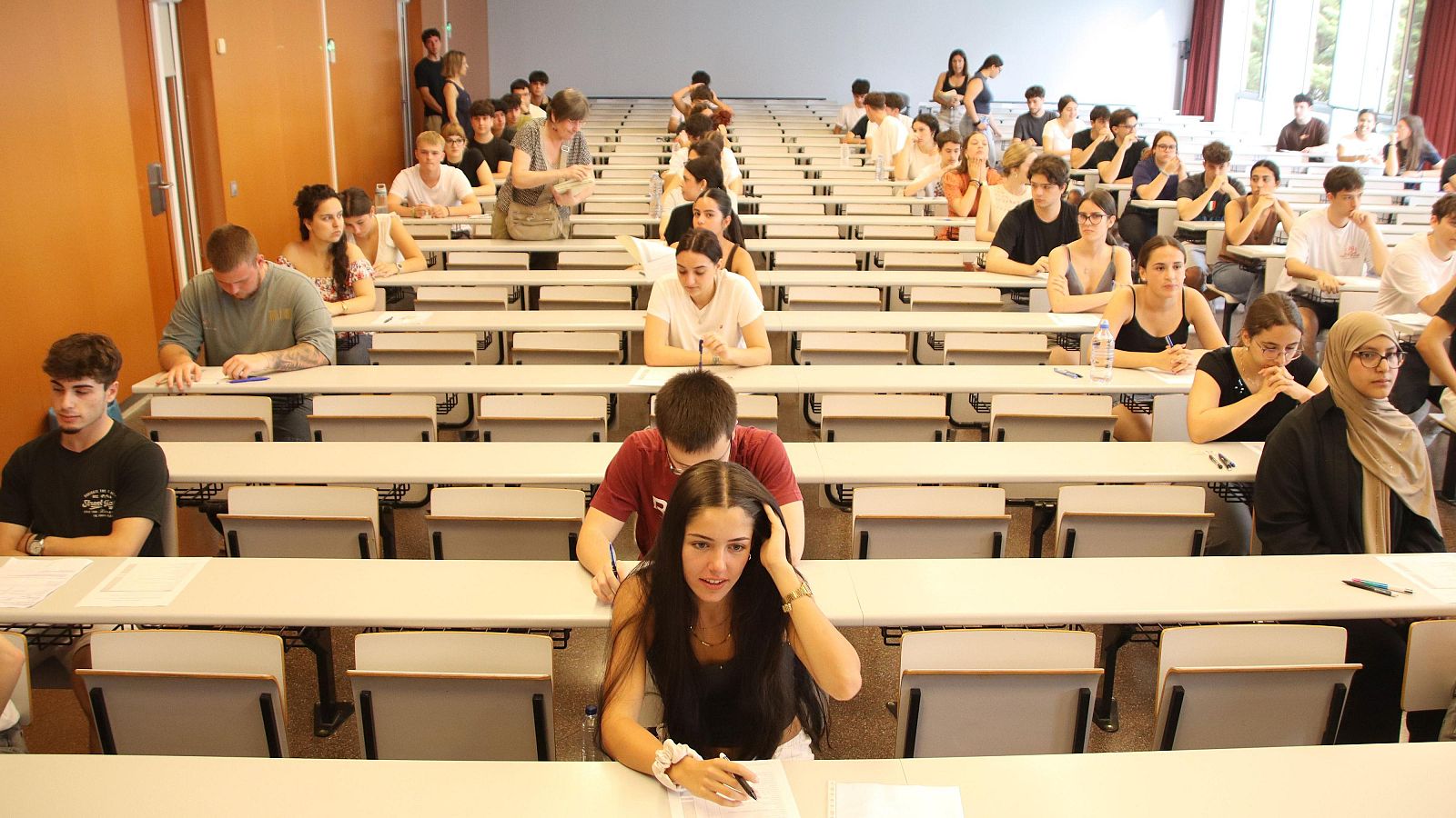 Aula de exámenes con numerosos estudiantes jóvenes concentrados en sus pupitres. Ambiente de seriedad y estudio.