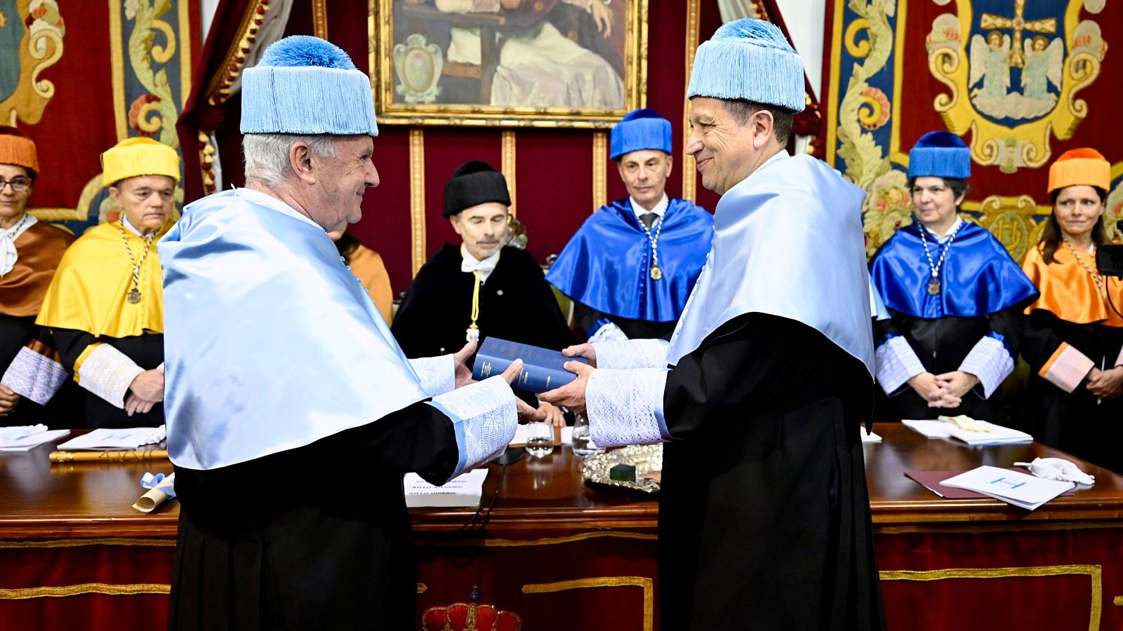 Víctor Manuel, con toga azul claro, recibe un libro de un académico en la Universidad de Oviedo durante su investidura como Doctor Honoris Causa. El evento se celebra en un salón adornado con tapices rojos y dorados.