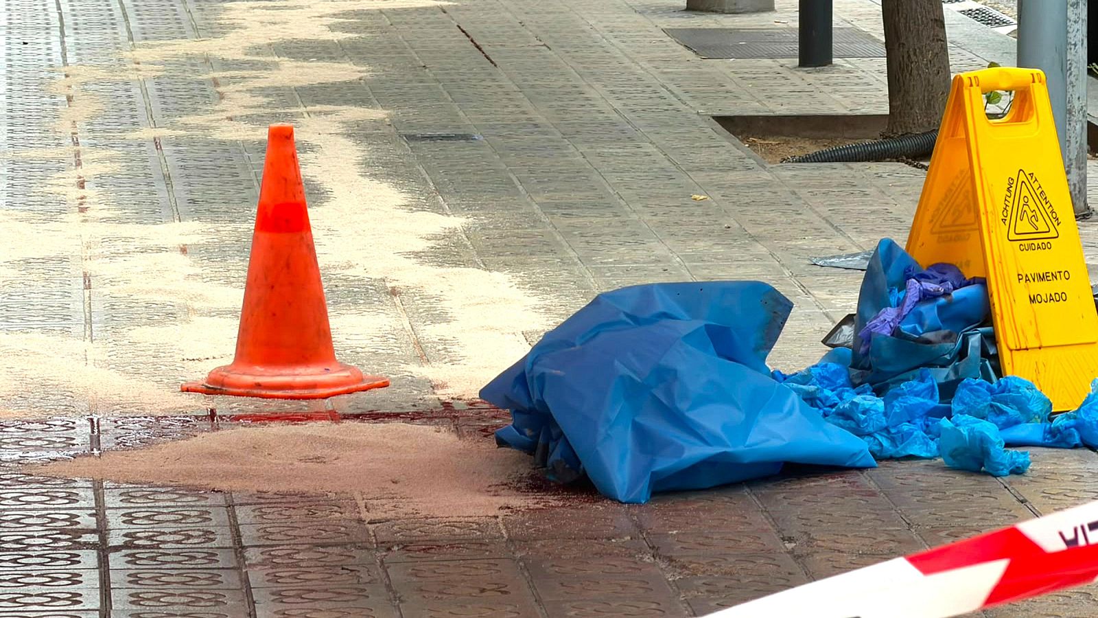 Escena urbana con cinta de policía, material absorbente marrón, plástico azul y señal de suelo mojado. El contexto sugiere un incidente en la calle Balmes de Barcelona.