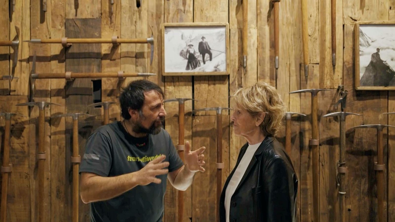 Álex Txikon y Mercedes Milá conversan en un escenario con piolets antiguos y fotos de montaña. Txikon gesticula, Milá escucha atentamente. El fondo evoca la historia del alpinismo.
